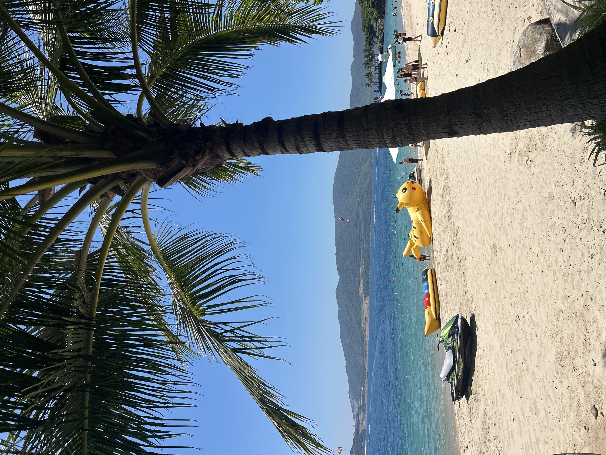 A vibrant tropical beach scene unfolds under a clear blue sky in Nam Nha Trang Ward, Vietnam. A large palm tree with dark, textured bark dominates the foreground, its fronds casting dappled shadows on the golden sand. To the left, a green and black Yamaha jet ski rests on the shore. Further down the beach, an inflatable yellow Pikachu toy lies on the sand, with a yellow and red inflatable banana boat positioned in the shallow turquoise water. Several people are visible on the beach and in the water, some wading, others relaxing, and a few appear to be participating in water activities. In the distance, lush green mountains form a scenic backdrop to the sparkling sea, and a parasail can be seen in the sky. White umbrellas and more inflatable toys are scattered along the shoreline. The overall atmosphere is one of a sunny, active, and recreational beach day.