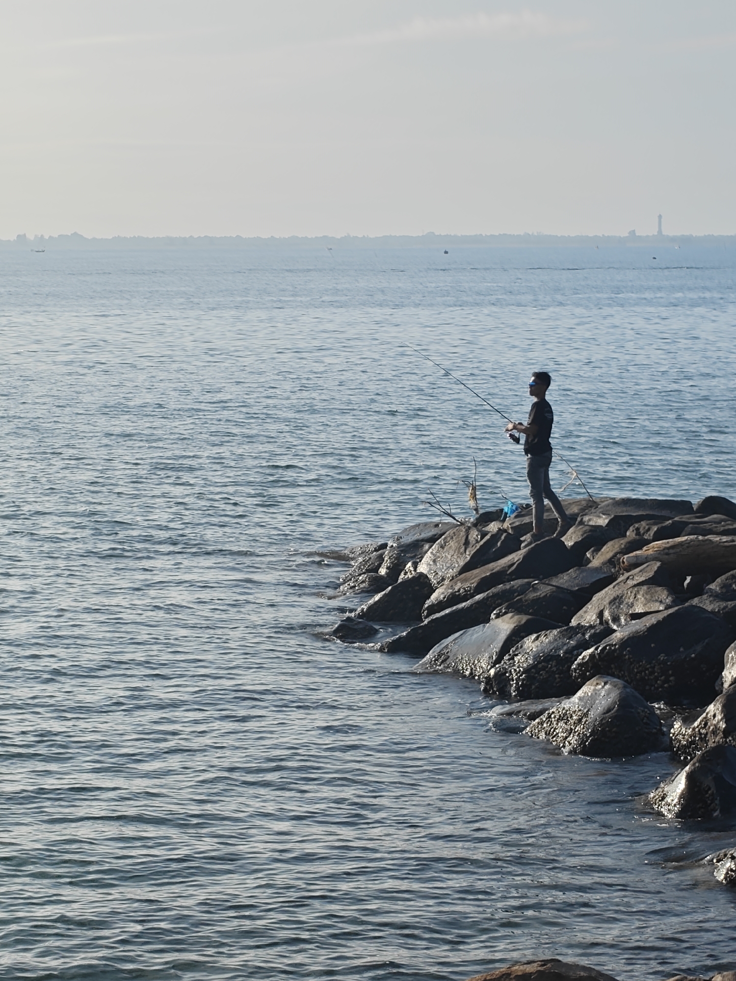 A lone fisherman stands on a rocky breakwater, angling in the sea off the coast of Lhokseumawe, Indonesia. The man wears sunglasses and dark clothing. The water is calm, with small waves lapping against the rocks. In the distance, the faint outline of buildings and a tall structure are visible. The sky is overcast and hazy.