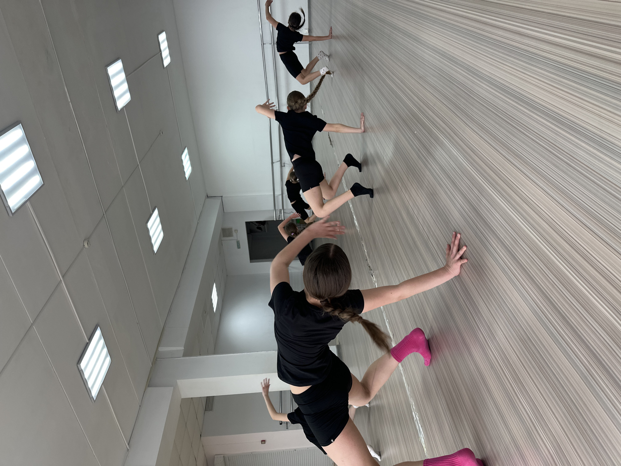 The image shows a dance studio, likely in Votkinsk, Russia, with several girls in black shirts and shorts practicing dance moves. They are positioned against what appears to be a mirror wall, giving the illusion of them being on a slanted surface. One girl in the foreground wears bright pink socks. The studio has multiple rectangular ceiling lights. The floor has a striped pattern. The scene is captured from a low angle.