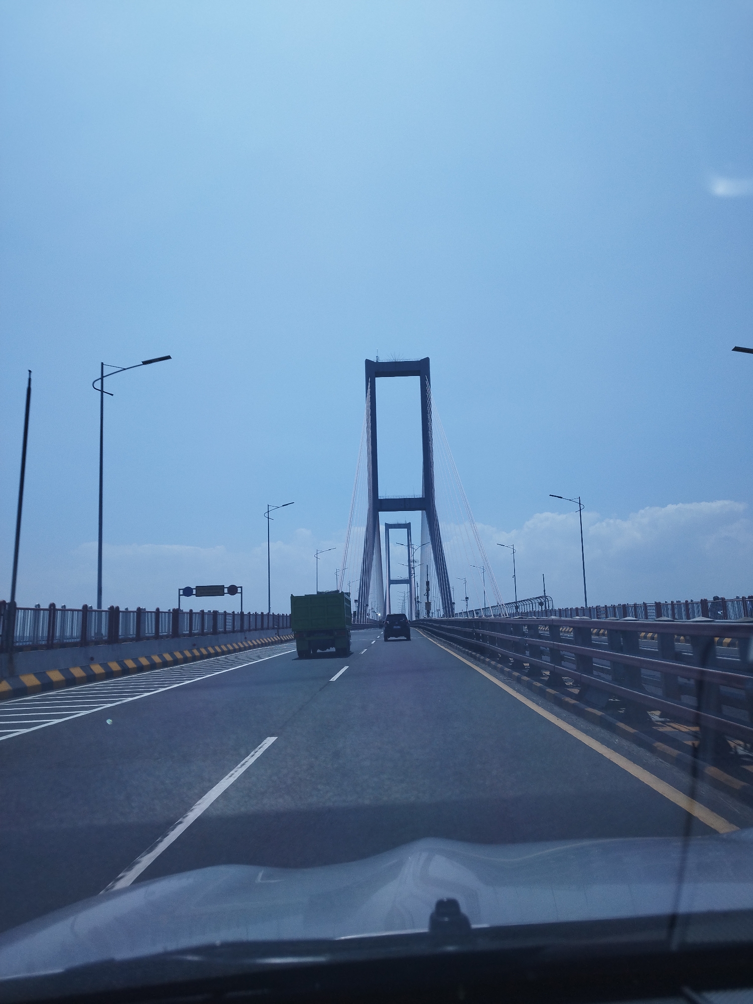 The image shows a view from the driver's seat of a vehicle traveling across a suspension bridge on a clear, overcast day. The bridge has multiple lanes, indicated by white lane markings. On the left side of the road, there is a railing with vertical bars and horizontal supports, topped with yellow and black striped markings. Streetlights are positioned along the left side of the bridge. In the distance, the prominent structure of the suspension bridge is visible, with tall, A-frame towers supporting the cables. Several vehicles are on the bridge, including a green truck in the right lane and a dark passenger car further ahead in the left lane. The sky is a pale blue with some faint white clouds. The location is identified as the Suramadu Bridge in Surabaya, Indonesia, not Pangpong.