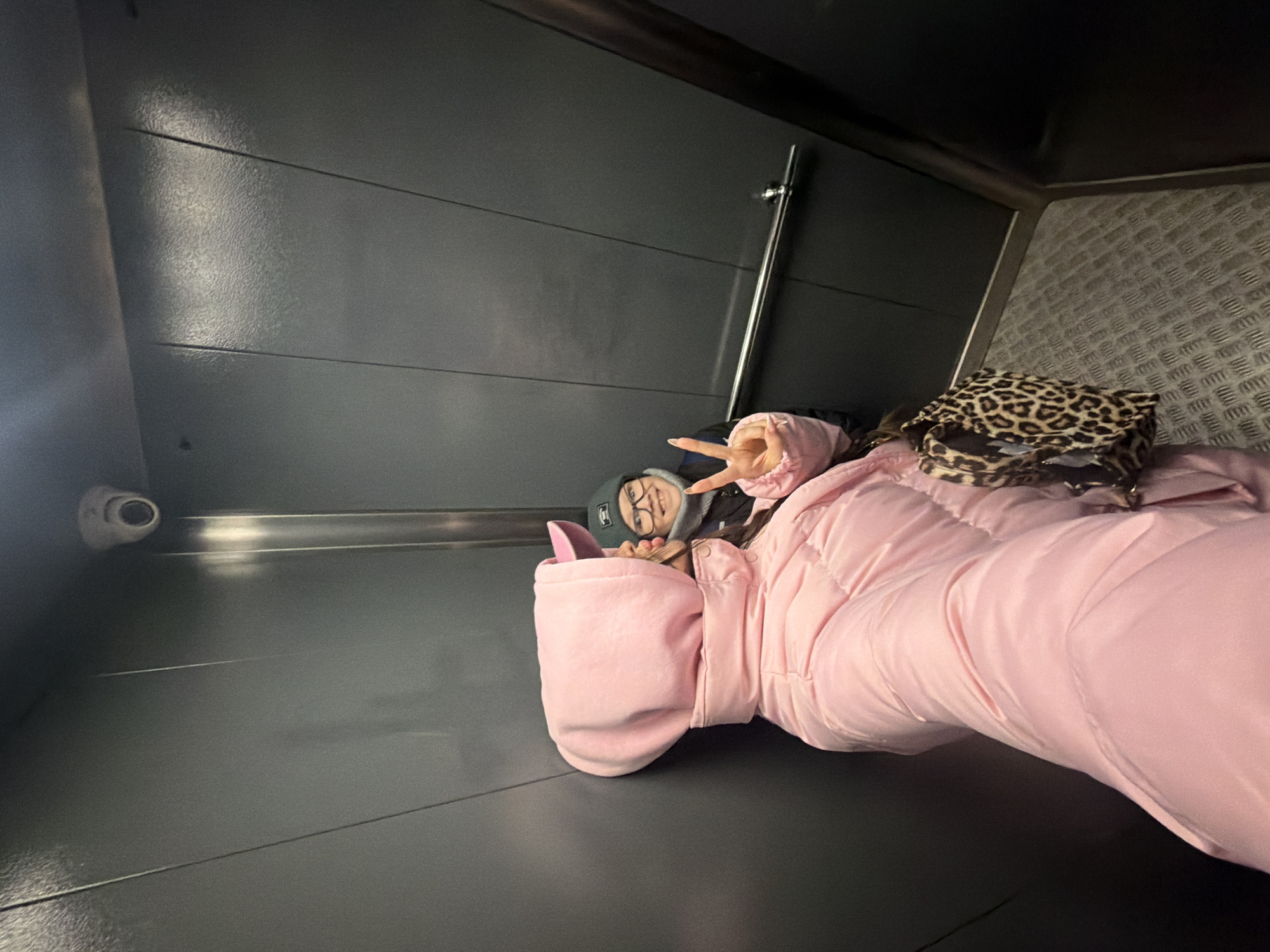 The image appears to be taken inside a lift or elevator, given the presence of the wall panels, handrail, and CCTV camera. A person wearing a pink puffer jacket and grey beanie is reclining on the floor, giving a peace sign. A leopard print bag rests beside them. The presence of metal diamond plate in the background suggests the lift might be in an industrial or service area. However, the information is insufficient to determine whether the location is in the city of Saint Petersburg, Russia.