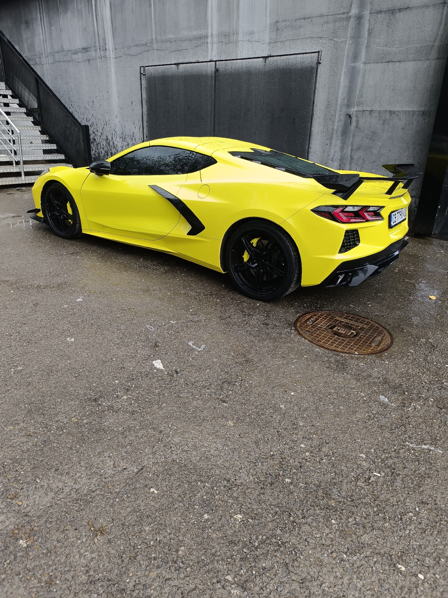 The image shows a yellow Chevrolet Corvette parked on an asphalt surface next to a concrete wall and a set of metal stairs. The car is facing towards the right with the rear of the vehicle in view. The car has black wheels, black side mirrors, a black rear spoiler, and black accents along the side. The asphalt appears wet and has some scattered debris. A circular manhole cover is located to the right of the rear of the car. The license plate on the car is visible, although the characters are not clearly legible. Based on the license plate format, the scene is likely in Sofia, Bulgaria.