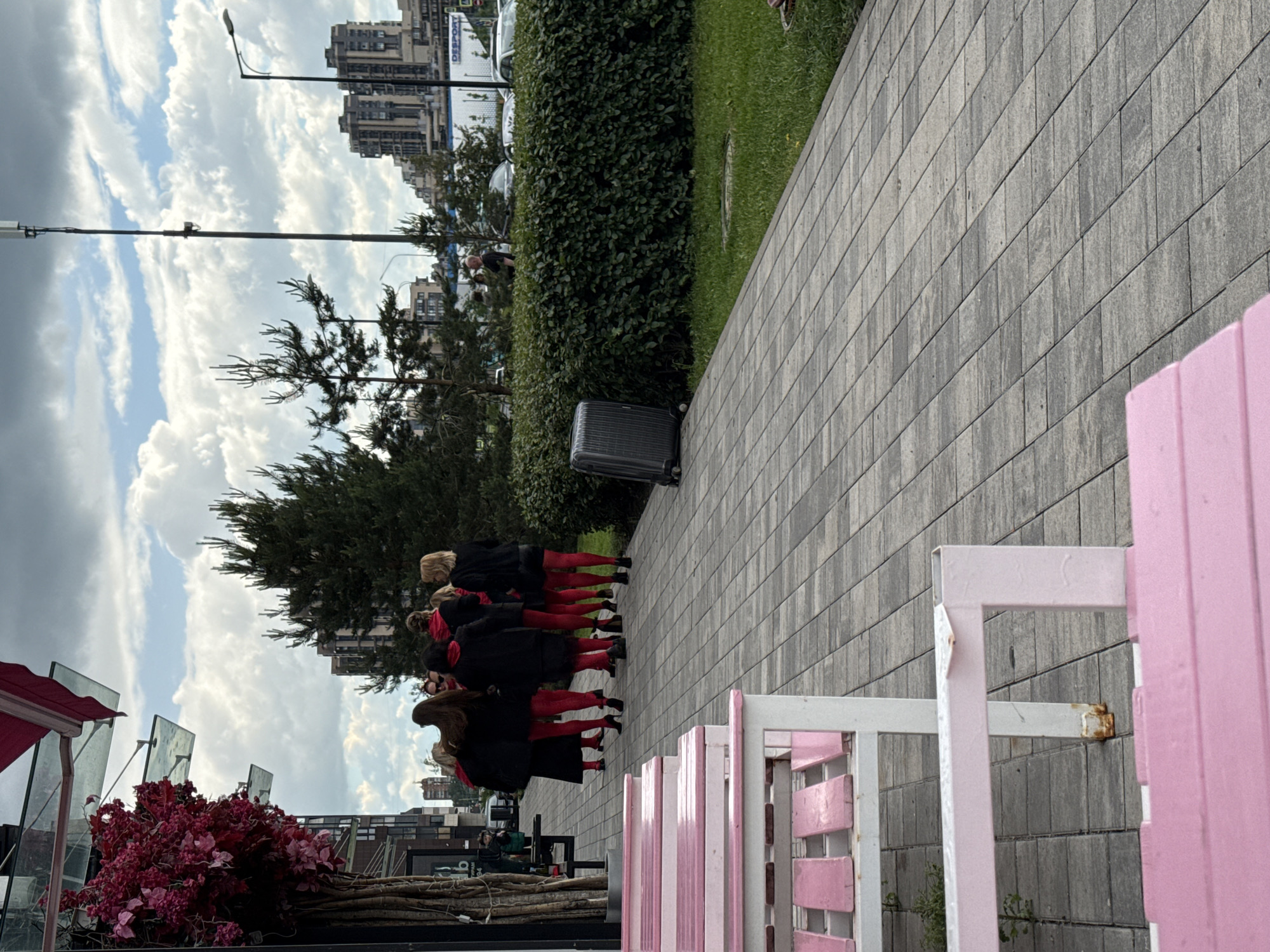 In an outdoor urban setting, a group of seven women, all wearing black tops and bright red tights, stand on a paved walkway. They are positioned in front of a dense green hedge and near tall coniferous trees. To the left, pink benches are visible, with a building featuring a large red awning and hanging greenery. In the background, modern high-rise buildings are silhouetted against a cloudy sky. A dark grey suitcase rests on the walkway near the hedge. The scene appears to be a street or public area in a city.