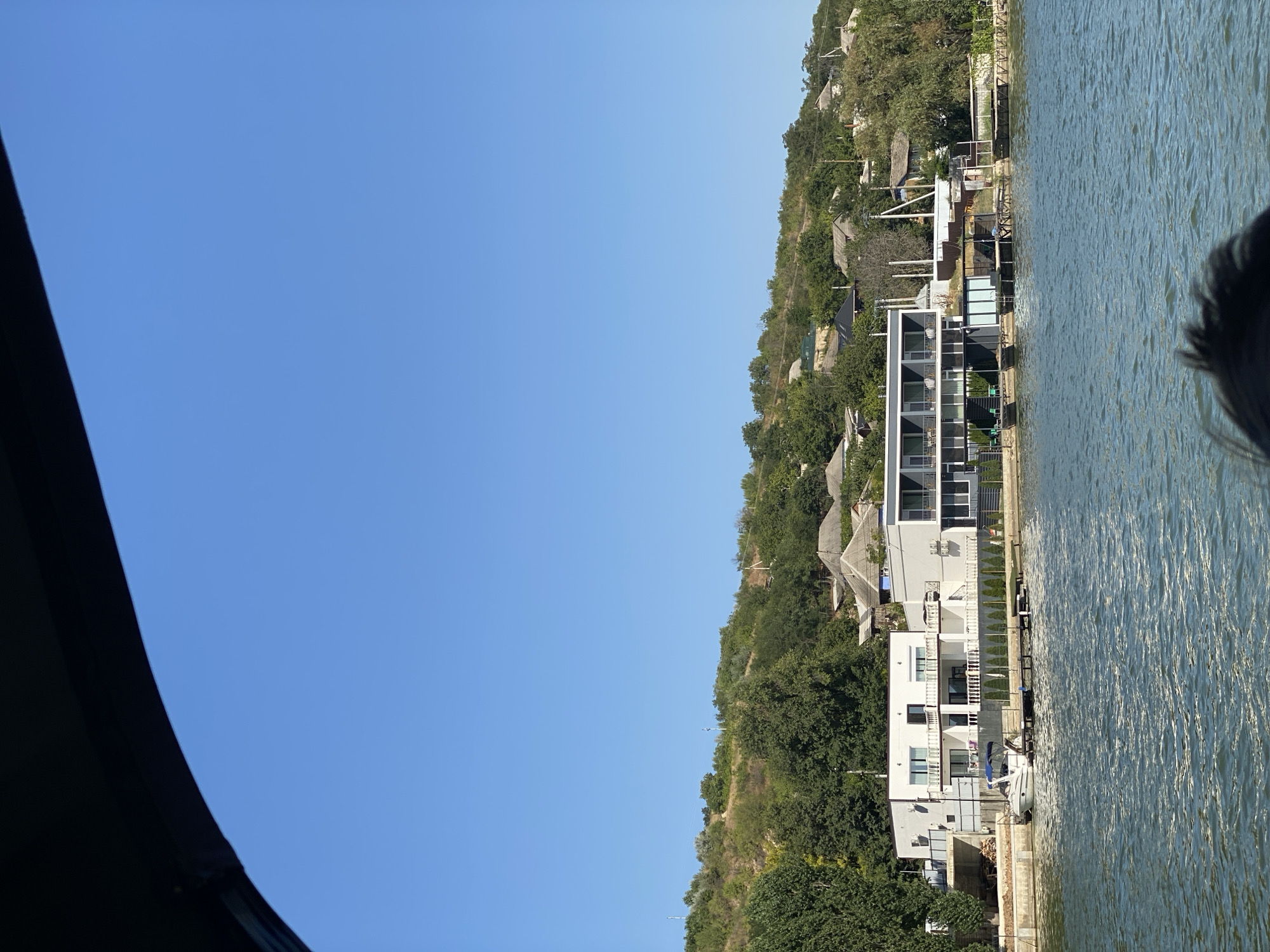 The image depicts a waterfront view in Molovata, Moldova, under a clear blue sky. In the foreground, a body of water with moderate ripples occupies the lower half of the frame. Along the shoreline, a modern, white, two-story building with balconies and large windows is visible, situated next to a contemporary grey building. A small white motorboat is docked at a pier in front of the white building. Several other structures, including houses with pitched roofs, are scattered on a tree-covered hillside rising behind the waterfront buildings. The overall scene suggests a peaceful, sunny day in a riverside community.