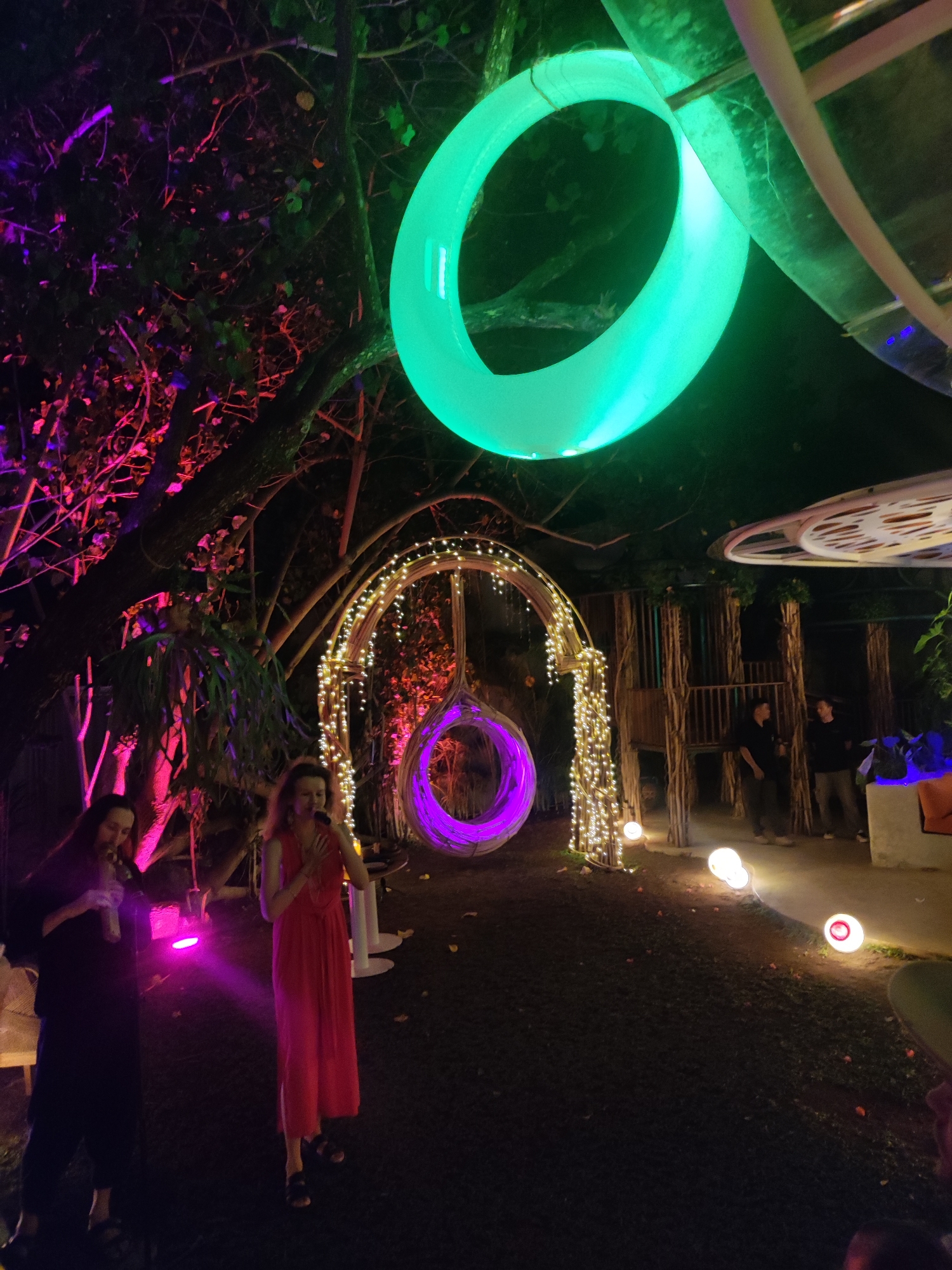 In a nocturnal outdoor setting, possibly a garden or park in North Kuta, Indonesia, illuminated by vibrant artificial lights, several individuals are present. Two women stand in the foreground; one on the left, dressed in dark attire, holds a drink. The woman on the right, wearing a long red dress, appears to be speaking or singing into a microphone. Behind them, a large archway adorned with twinkling fairy lights frames a purple, circular hammock-like swing. Further back, a wooden structure resembling a treehouse is visible, with two men standing near its entrance.
The scene is dominated by large, glowing rings of light – one a bright green overhead, and another, purple, is the swinging chair. The trees are lit with pink and purple lights, creating a festive and somewhat mystical atmosphere. Small, round lights are scattered along the pathway. The time of day is clearly night, suggested by the darkness and the reliance on artificial illumination. The overall mood appears to be celebratory or social, with the lighting contributing to a magical, immersive experience. No visible text is present in the image.