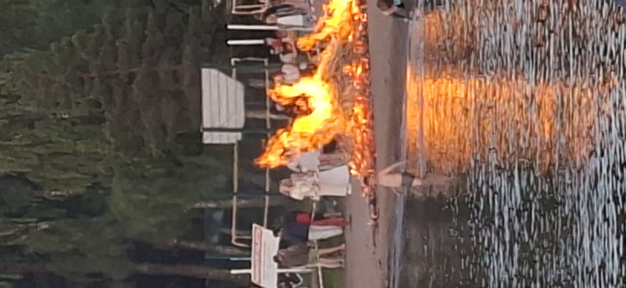 A bonfire is burning on a sandy area next to a body of water. A young boy wearing dark shorts is standing in the water, facing the fire. The fire is large and consists of logs and branches, producing bright orange and yellow flames. Several people are gathered on the sandy area behind the fire, some are standing and watching, while others appear to be socializing. A few trees are visible in the background. The scene appears to be taking place in the evening. This is a scene from the city of Salpakangas, Finland.