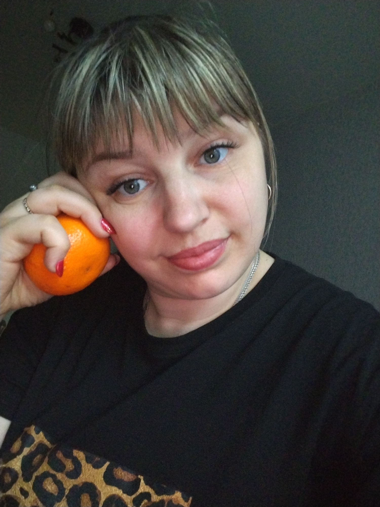 A woman is posing for a selfie. She has fair skin, light brown hair with blonde highlights, and blue eyes. She is wearing a black t-shirt with a leopard print design visible at the bottom of the frame. The woman is holding an orange against her face with her left hand, where she has a ring and red nail polish. She is wearing a silver necklace and hoop earrings. The background is a dark, indistinct wall. The location is not identifiable as Yaroslavl, Russia, based on the visual information provided.