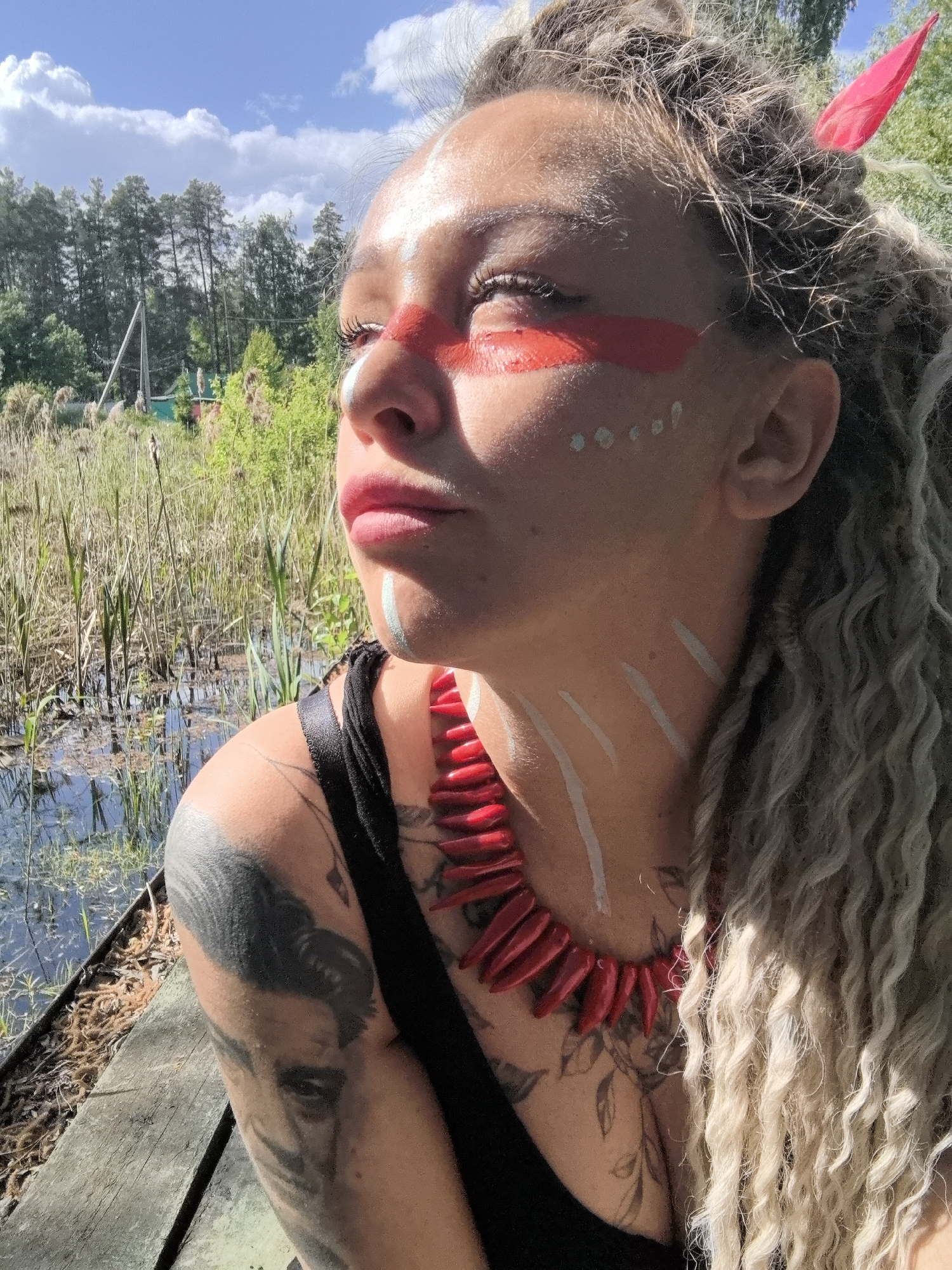 A woman with light-colored dreadlocks and tribal-inspired face and body paint is sitting near a body of water. She has a red stripe across her cheekbones, white dots near her ear, and white stripes on her neck. She wears a red, tooth-shaped necklace and a red feather adornment in her hair. Visible tattoos are on her arm and chest. The woman is seated on a wooden structure. Behind her, there is a body of water with tall reeds and a forest in the background. The scene is illuminated by sunlight, casting shadows and highlights. The location is Noviy, Russia.