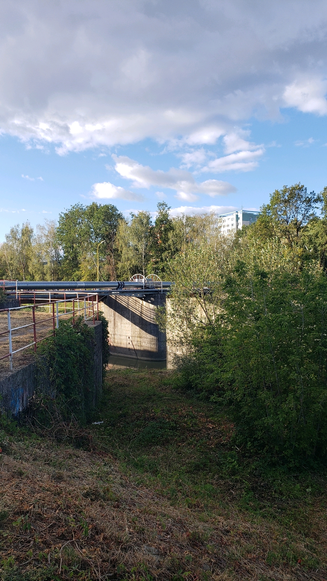 A medium shot captures a canal lock structure with a large pipe overhead, situated in a vegetated area with trees and a building in the background. The sky is partly cloudy, with a mix of blue sky and white and gray clouds. The canal lock is constructed of concrete and features a metal railing along its edge. The large pipe, which appears to be part of an industrial infrastructure, runs horizontally across the top of the lock. Two white, gear-like mechanisms are visible on the pipe. Dense green foliage and trees surround the canal lock, and the ground is covered with grass and fallen leaves. In the distance, a modern, multi-story building with white and blue accents is visible, suggesting an urban or suburban setting. The lighting suggests it is daytime, with the sun casting shadows. The overall scene depicts a functional piece of infrastructure integrated into a natural or semi-natural environment. This is likely the canal lock on the Torysa River in Prešov, Slovakia, near the city center.