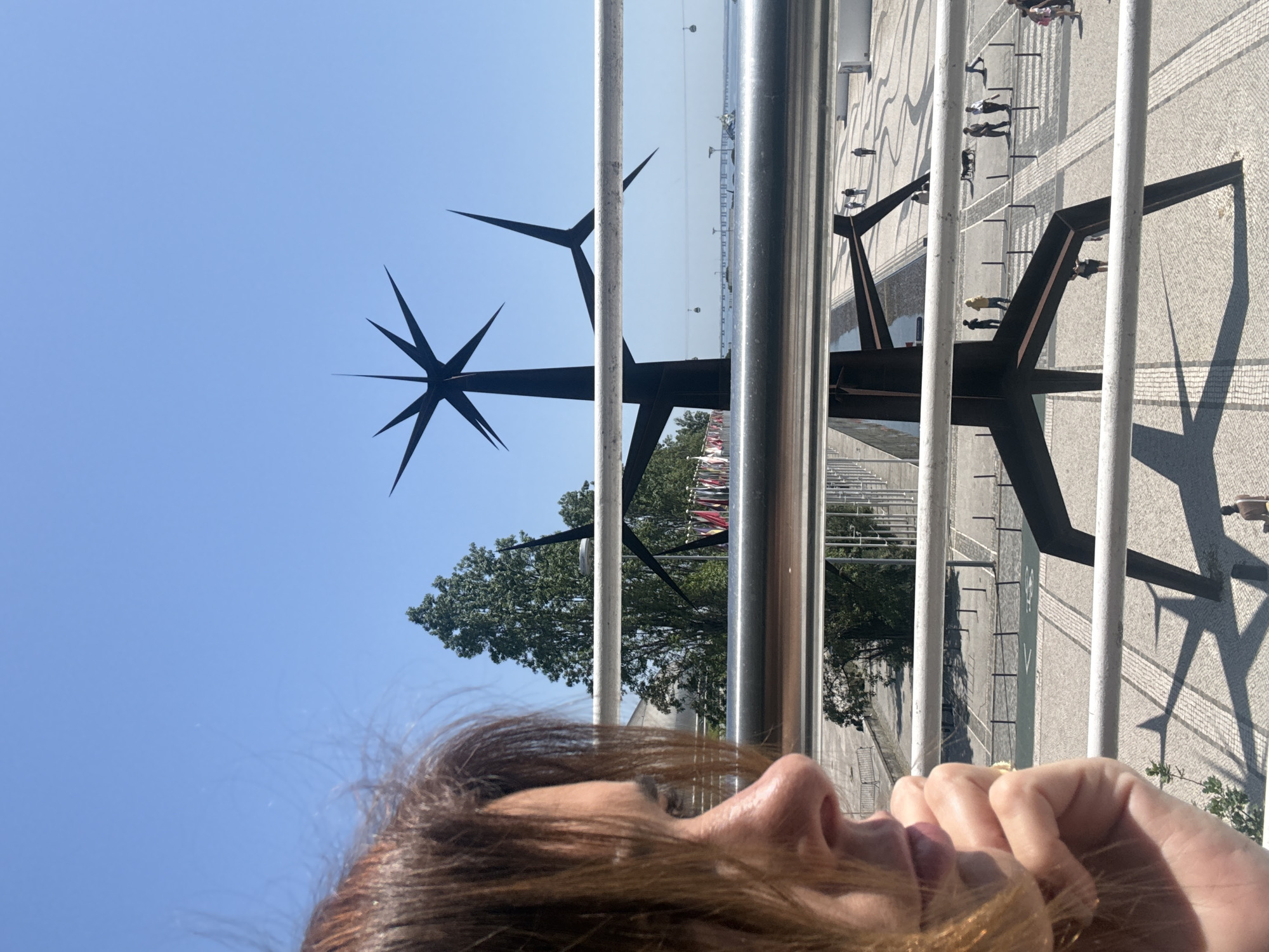A woman is viewing the Monument to the Discoveries (Padrão dos Descobrimentos) in Lisbon, Portugal, through a metal railing. The monument, featuring prominent spike-like elements, stands against a clear blue sky. In the background, the Tagus River and the patterned pavement of the plaza where the monument is located are visible. People are walking on the plaza. The image is taken at an odd angle, with the woman's face and hair in the foreground.