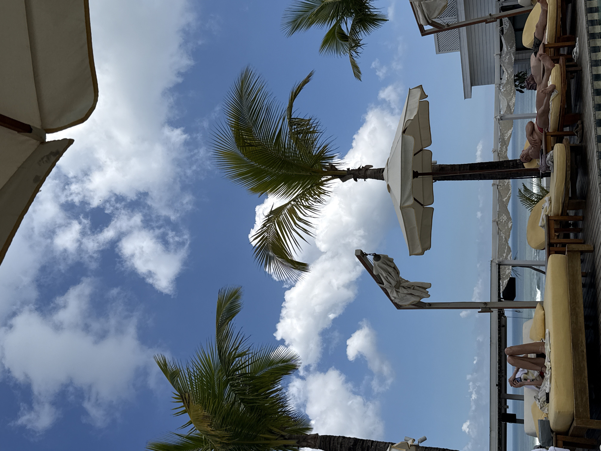 The image depicts a beach scene in North Kuta, Indonesia. The view is angled upwards, showcasing palm trees against a blue sky with scattered white clouds. Several white sun umbrellas are visible, some extended and providing shade, others closed. Lounge chairs are arranged on a wooden deck, occupied by people relaxing. The ocean is visible in the background.