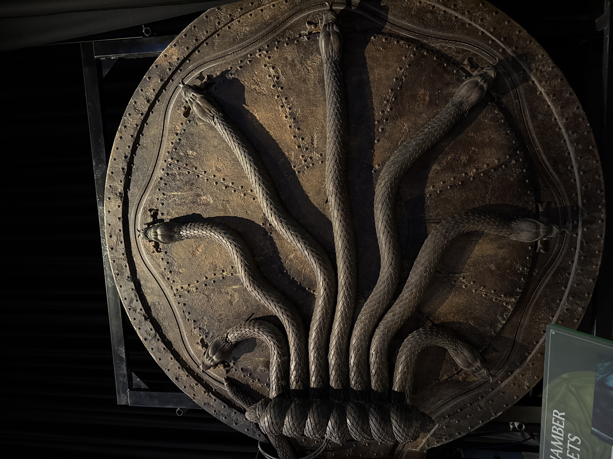 The image shows a large, circular, weathered metal door decorated with multiple sculpted snakes. The snakes are dark and appear to be intertwined, with their heads facing outwards or coiled around the door's surface. The door itself is a dark, rusty brown color and is covered in numerous small rivets. It is mounted within a black metal frame and is set against a black backdrop. In the lower left corner, a green sign with white text is partially visible, with the words "HAMBER" and "ETS" clearly legible. This object is likely on display at Warner Bros. Studio Tour London – The Making of Harry Potter in Watford, United Kingdom, given its appearance and the visible text which suggests it is related to the "Chamber of Secrets" from the Harry Potter series. There are no people visible in the image, and the scene appears to be a still display.