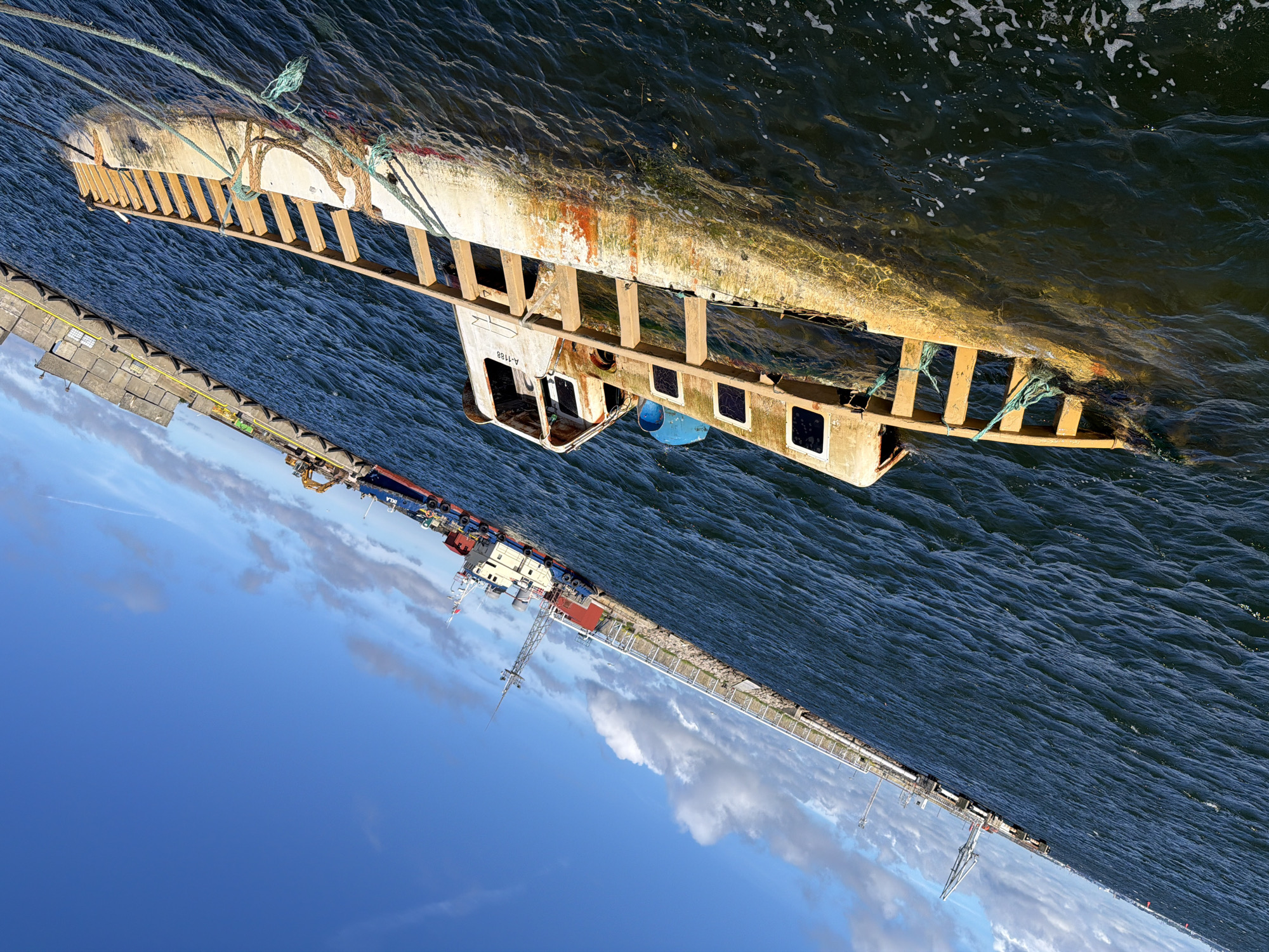 A sunken vessel, partially submerged in dark blue water, lists to its port side. The vessel appears to be a small fishing boat or tug, with a weathered white and orange hull. A wooden railing runs along its deck. The vessel is tethered by ropes to an unseen point. In the background, a long pier juts out into the water, lined with industrial equipment including cranes and what appear to be shipping containers. Further in the distance, other vessels are docked or moored. The sky above is a clear, bright blue with scattered white clouds. The scene appears to be a harbor or industrial port area, likely in the city of Viimsi, Estonia, given the context. The water is moderately choppy.