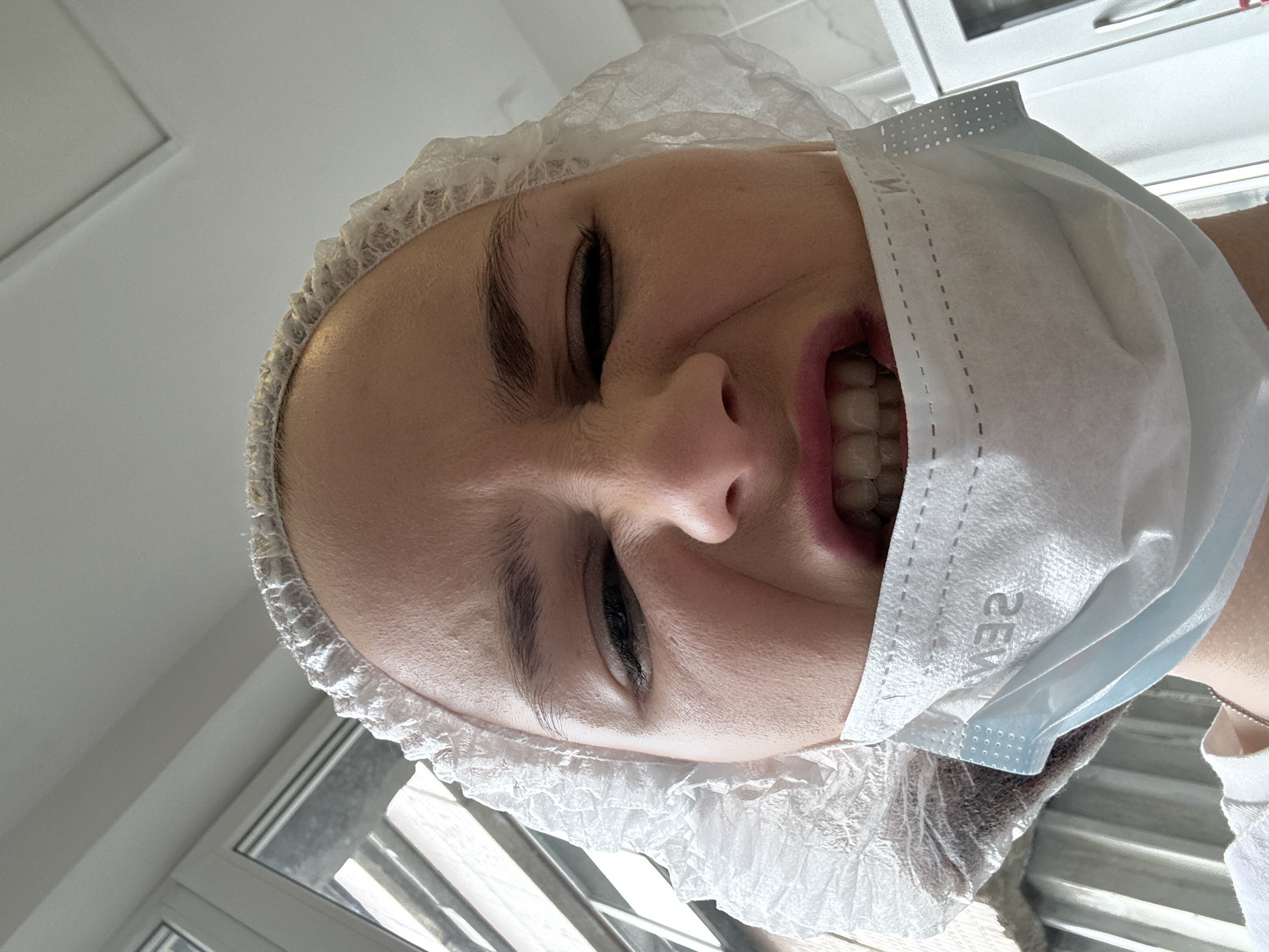 A fair-skinned woman wearing a white disposable surgical cap and light blue medical face mask smiles, squinting her eyes. The mask obscures part of her face and displays a reversed inscription, possibly indicating the manufacturer. Her teeth are visible. The location might be a medical or cosmetic setting, potentially in Saint Petersburg, Russia, as per the request, although there's no definitive visual evidence confirming the location. A window and cabinet are partially visible in the background.