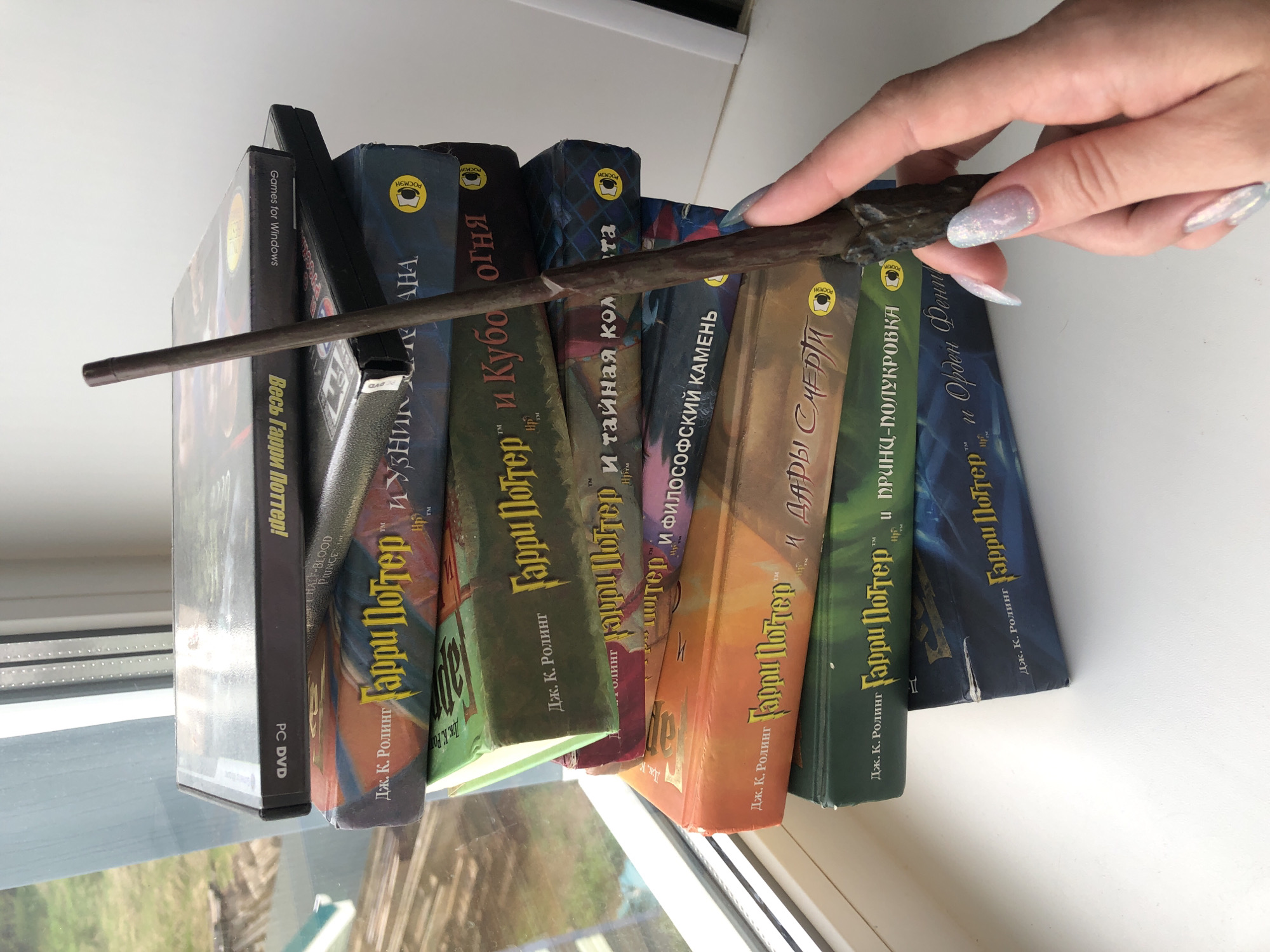 A photograph taken indoors shows a collection of Harry Potter books and a video game, arranged in a slightly slanted row. The books are the Russian editions, authored by J.K. Rowling, and include "Harry Potter and the Philosopher's Stone," "Harry Potter and the Chamber of Secrets," "Harry Potter and the Order of the Phoenix," "Harry Potter and the Half-Blood Prince," and "Harry Potter and the Goblet of Fire." Adjacent to the books is a DVD game titled "All Harry Potter." A hand with long, manicured nails holds a dark-colored wand horizontally across the books. The scene is situated near a window, as evidenced by the natural light and the glimpse of greenery visible outside. The books display the "РОСМЭН" logo, indicating they were published in Russia. The setting appears to be a windowsill in Noviy, Russia.