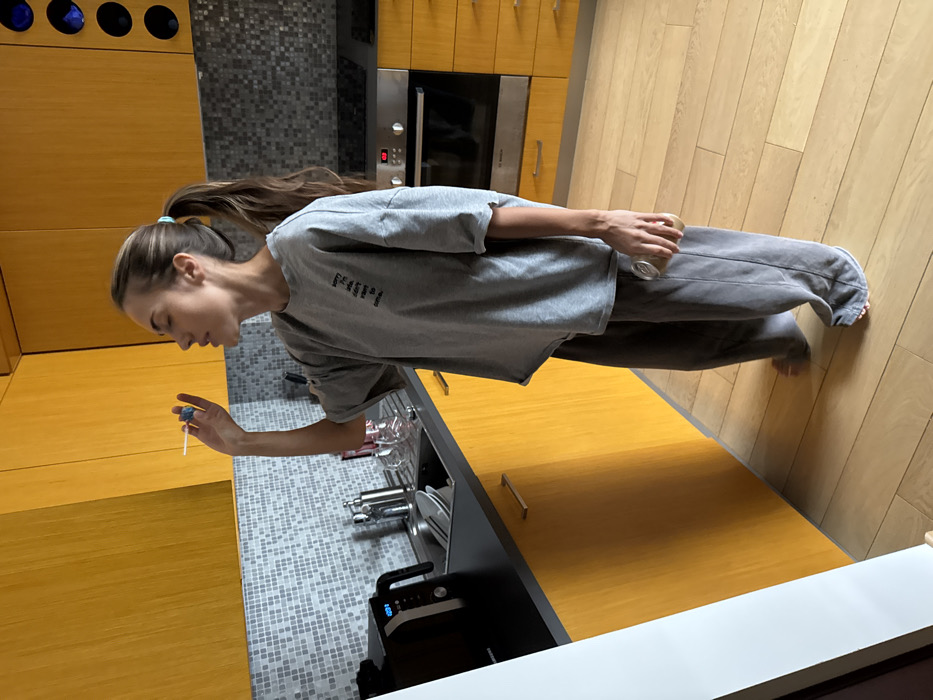 A young adult female is standing barefoot in a modern kitchen, holding a blue and white lollipop to her mouth. She is dressed in a loose-fitting gray short-sleeved t-shirt with dark text on the chest and dark gray wide-leg sweatpants. Her brown hair is tied back in a high ponytail. In her left hand, held loosely at her side, she holds a small, clear glass bottle with a brown label.
The kitchen features yellow wooden upper and lower cabinets with minimalist handles and a dark gray countertop. The backsplash is composed of small, light gray square mosaic tiles. A stainless steel sink with a faucet is visible, containing several dishes. To the left of the sink, a black microwave oven displays "12:01". To the right, a stainless steel oven and stovetop unit is integrated into the cabinetry, beneath a wall-mounted wine rack with blue bottles. The floor is light-colored wooden laminate. The scene takes place in a residence in Warsaw, Poland.