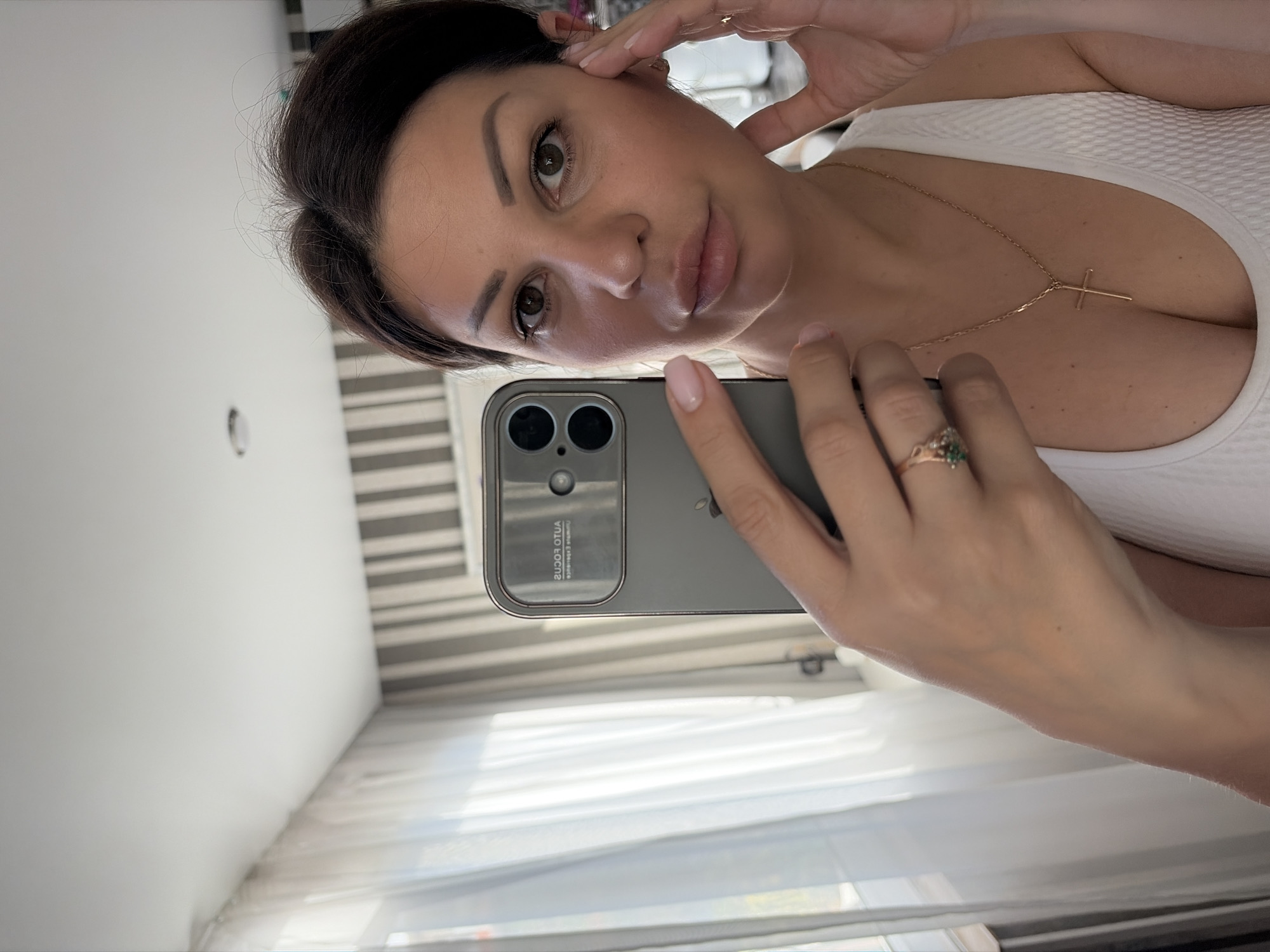A woman is taking a selfie in a room with striped wallpaper and sheer curtains. She has dark hair and is wearing a white top with a cross necklace. She is also wearing a ring with a green stone on her left hand. The phone in her hand shows the text "AUTOFOCUS" and "ULTRA WIDE". The image does not provide any information about the city of Sochi, Russia, or any other specific location.
