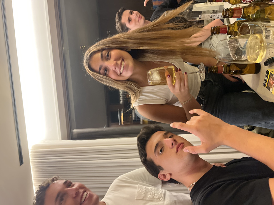 This image depicts an indoor social gathering involving four young adults at a residence in Lordelo do Ouro e Massarelos, Portugal.
A female with long, light brown hair, wearing a white v-neck top and dark jeans, smiles directly at the camera. She holds a stemmed glass containing an amber liquid with ice in her right hand; her fingernails are painted dark red. To her left, a male with dark hair, wearing a black t-shirt and a silver chain necklace, looks at the camera while making a "devil horns" hand gesture with his right hand. Partially visible on the far left, another male with light brown curly hair, wearing a white t-shirt, smiles towards the left. On the right, a fourth male, wearing a dark top, is partially visible, smiling towards the right.
The group is gathered around a white table laden with beverages. Visible items include several empty and partially filled amber glass bottles, identifiable as Super Bock beer. A clear glass bottle, likely containing spirits, and a wine glass with a light-colored liquid are also present. A yellow and white Marlboro cigarette pack and a small black cylindrical object, possibly an e-cigarette, are on the table surface. The background features a large window or glass door with light-colored curtains to its left, revealing a dimly lit interior or exterior space beyond. Recessed lighting fixtures are visible on the ceiling.