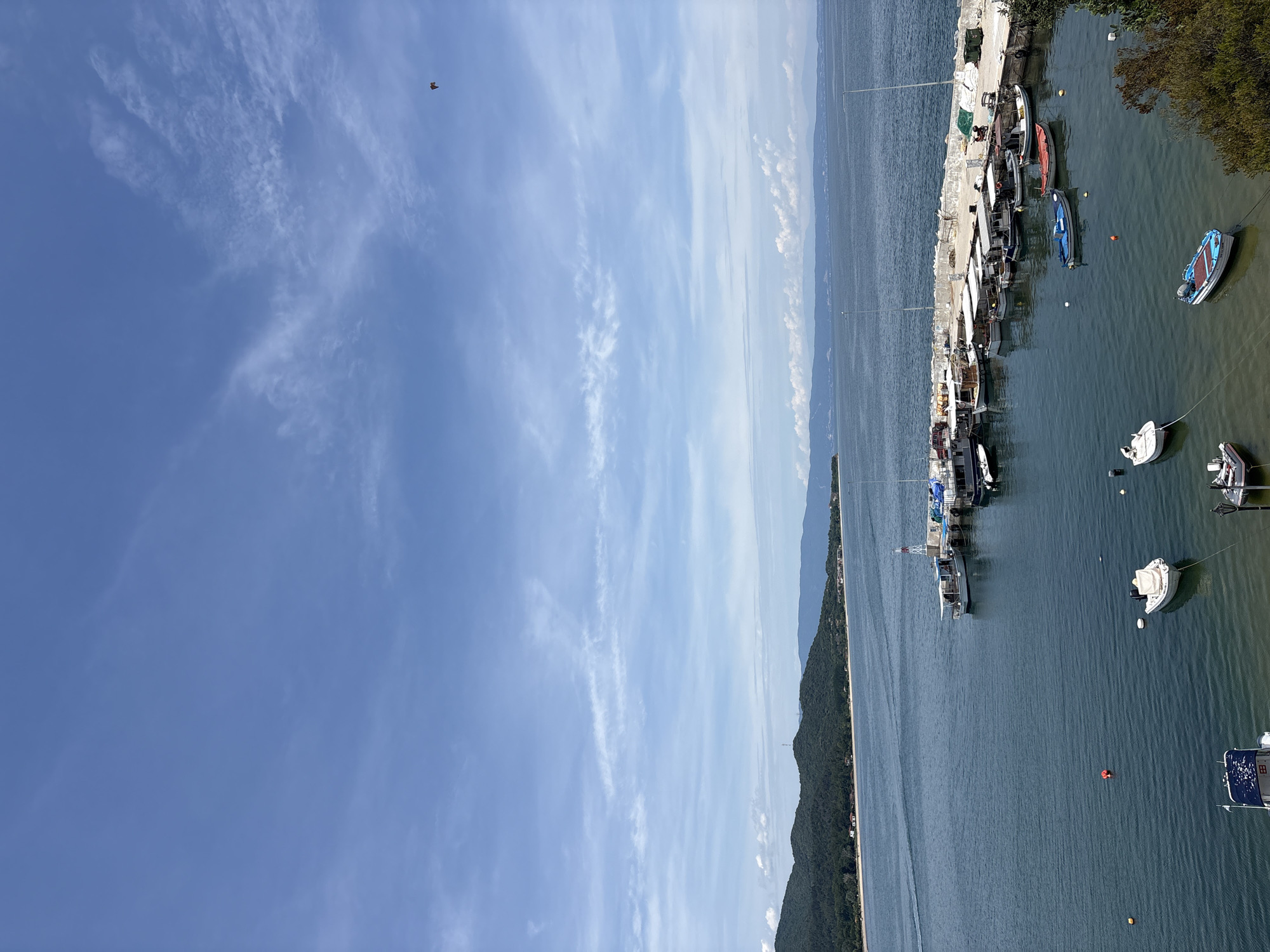 This is an aerial, high-angle view of a harbor in Olympiada, Greece, on a sunny day with a blue sky and scattered white clouds. The water is a deep blue, reflecting the sky, and has gentle ripples. A long, concrete pier juts out into the water, lined with numerous small to medium-sized fishing boats docked side-by-side. These boats are mostly white, with some featuring dark hulls or canopies. Several smaller vessels are moored in the calmer waters closer to the shore. There is a lighthouse structure on the pier, and various containers and equipment are visible along the pier's edge. In the distance, a lush, green, tree-covered mountain range forms the coastline, with some buildings scattered along its base and further inland. The sea extends to the horizon, where it meets a hazy, layered mountain landscape under the sky.