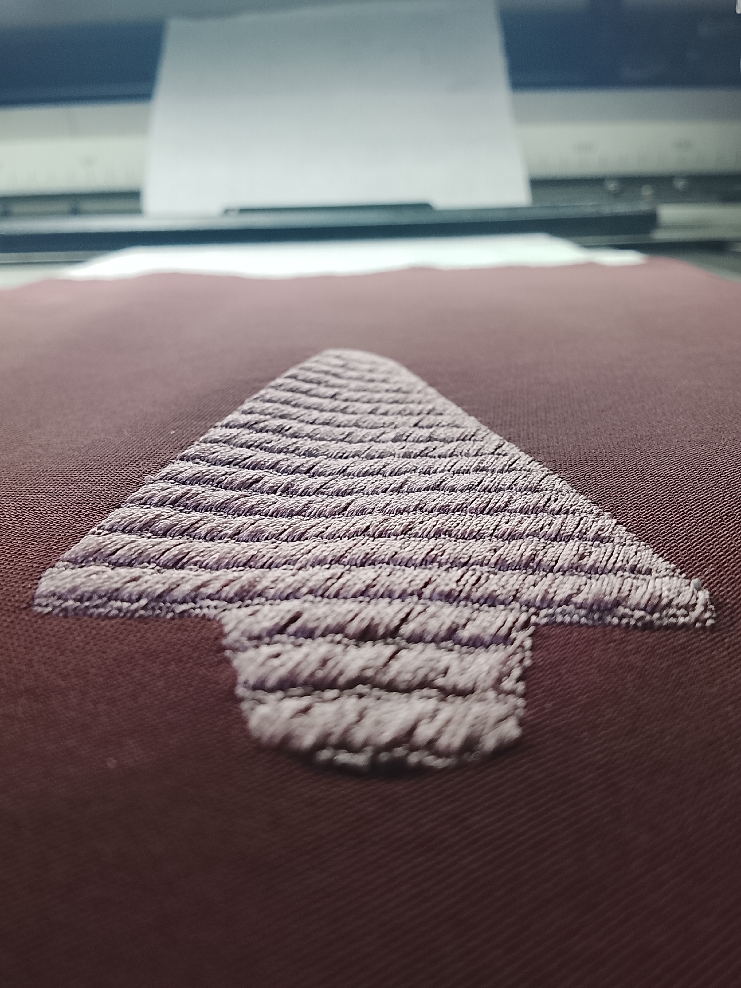 The image shows a close-up of a piece of fabric with an embroidered arrow pointing upwards. The fabric is a deep maroon color, and the embroidery is a lighter shade of purple or lavender. The embroidery is detailed, with visible threads creating a textured pattern within the arrow shape. The background is blurred and appears to be a machine, possibly an embroidery or printing machine, with a white sheet of paper or material being fed into it. The lighting suggests an indoor setting. There is no visible indication of people, actions, or interactions. The image is a still shot focused on the embroidered detail and the machinery. The location of Rumuepirikom, Nigeria is not discernible from the visual content of the image.