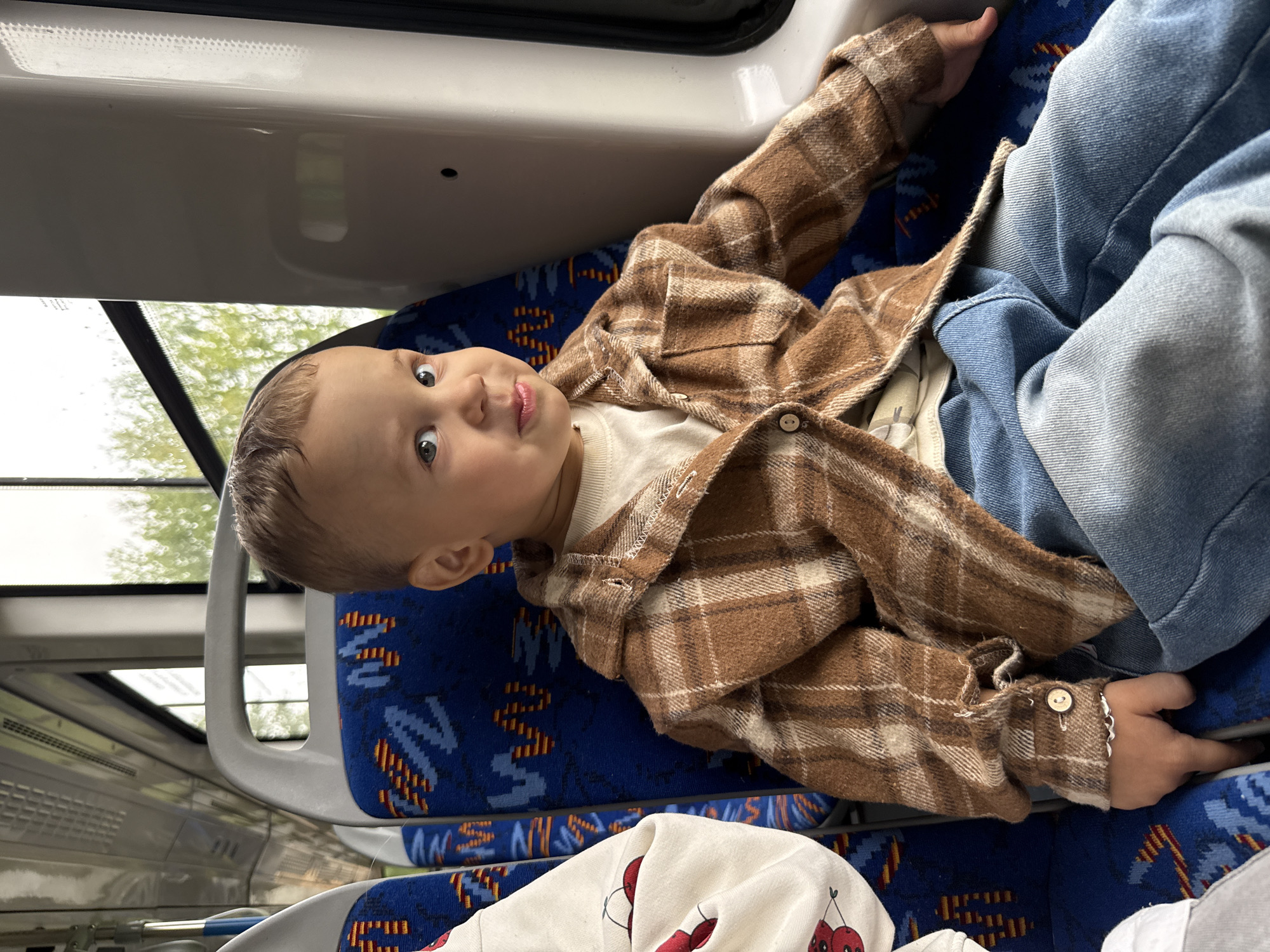In a bus, a young boy with light brown hair and green eyes is seated on a blue and red patterned seat. He is wearing a brown plaid shirt over a white t-shirt and blue jeans. The boy is looking towards the camera with his mouth slightly open, sticking out his tongue. To the left of the boy, a white garment with a cherry pattern is visible. The bus interior shows overhead compartments, windows with greenery visible outside, and adjacent seats. The location is stated to be the city of Rusanovo, Russia.