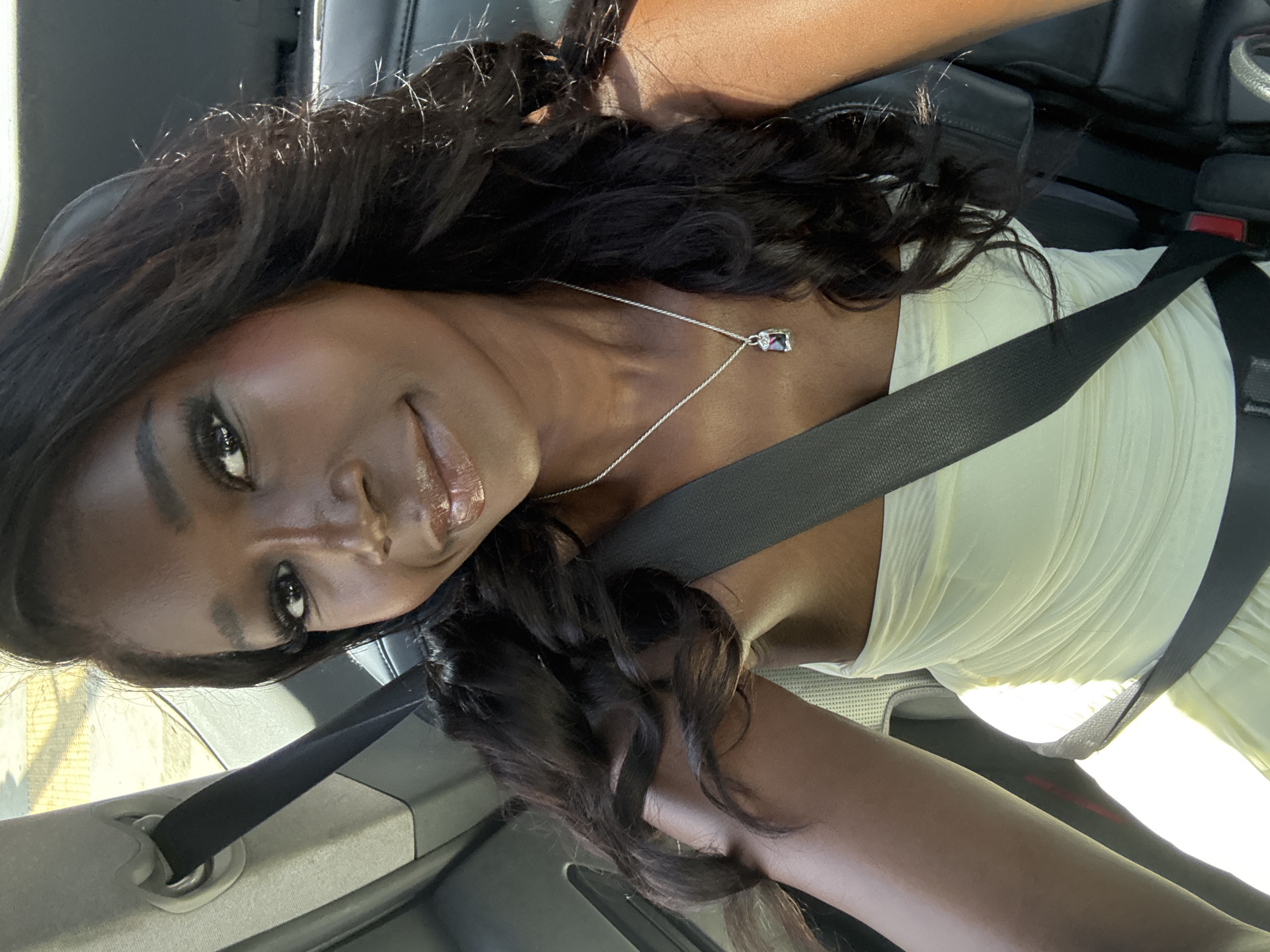 A dark-skinned woman is visible in a close-up, tilted shot taken inside a vehicle, likely a car, in Los Angeles, California. She is wearing a light-colored, possibly white or off-white, top and a seatbelt. She has long, dark, wavy hair and is wearing a delicate necklace with a small pendant. The interior of the car appears dark. The exact street address or location within Los Angeles is not discernible from the image.