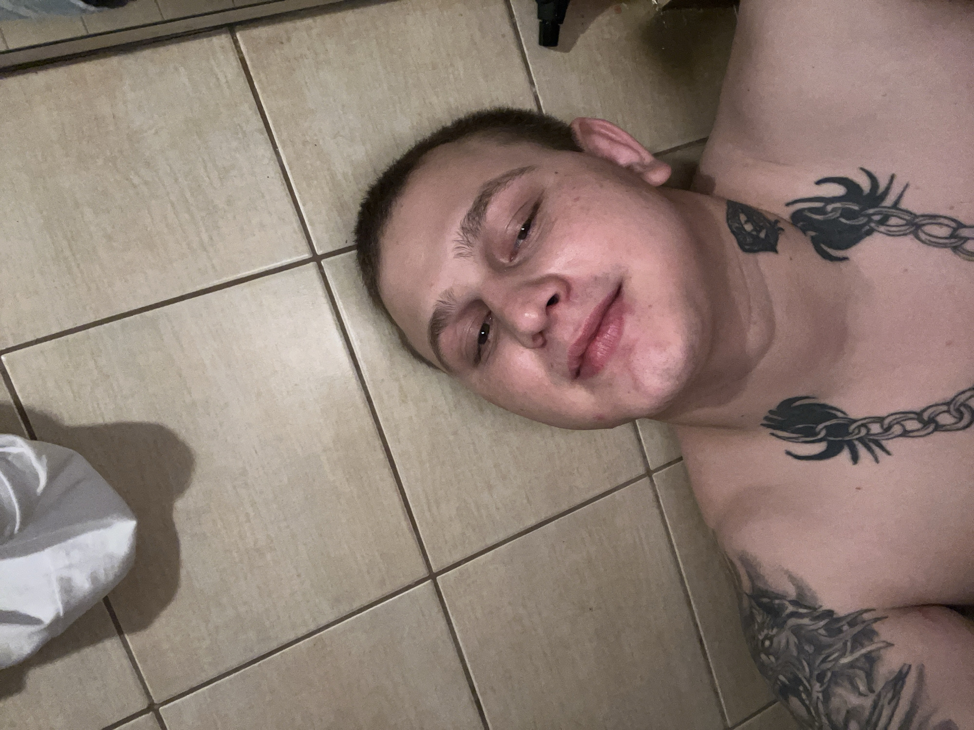 A person is lying on a tiled floor, looking up at the camera. The person has short hair and tattoos on their chest and arm. The floor tiles are light brown and laid in a grid pattern. There is a white cloth in the upper left corner of the image, casting a shadow. The scene is consistent with an indoor location, such as a bathroom or hallway, with tiled flooring. No specific location in Pilsen, Czechia, can be determined from the image.