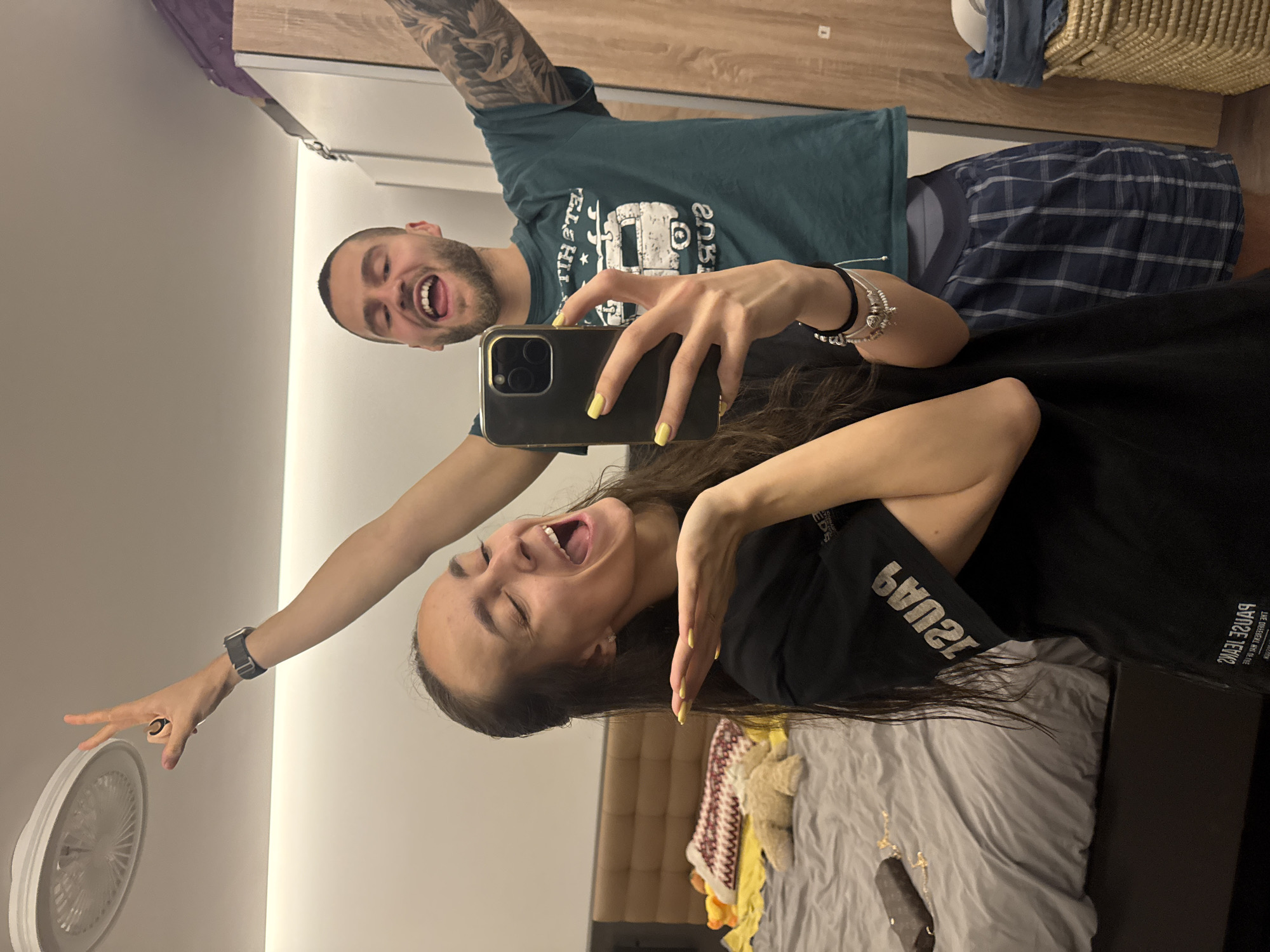 A playful mirror selfie captures a man and a woman in what appears to be a bedroom setting in Sofia, Bulgaria. The woman, positioned in the foreground, is holding a smartphone with a black and gold case to take the picture. She has long dark hair, bright yellow nail polish, and is wearing a black t-shirt with the mirrored text "PAUSE JEANS" (and smaller text below it) visible. Her left arm is extended upwards towards a partially visible ceiling fan, and she is wearing a black smart watch and silver bracelets on her wrists.
Both individuals are caught mid-laughter, with wide-open mouths and joyful expressions, indicating a lighthearted and happy moment. Reflected in the mirror behind the woman is the man, who has a beard, short hair, and visible tattoos on his left arm. He is wearing a dark green t-shirt featuring a graphic that appears to be a camper van and some mirrored text, where "THE BEST" is somewhat discernible. He is also wearing blue and white plaid pajama bottoms.
The background details include a bed with grey bedding, a brown headboard, a wooden wardrobe or closet, and a woven basket. The indoor lighting suggests the photo was taken in the evening or night.
