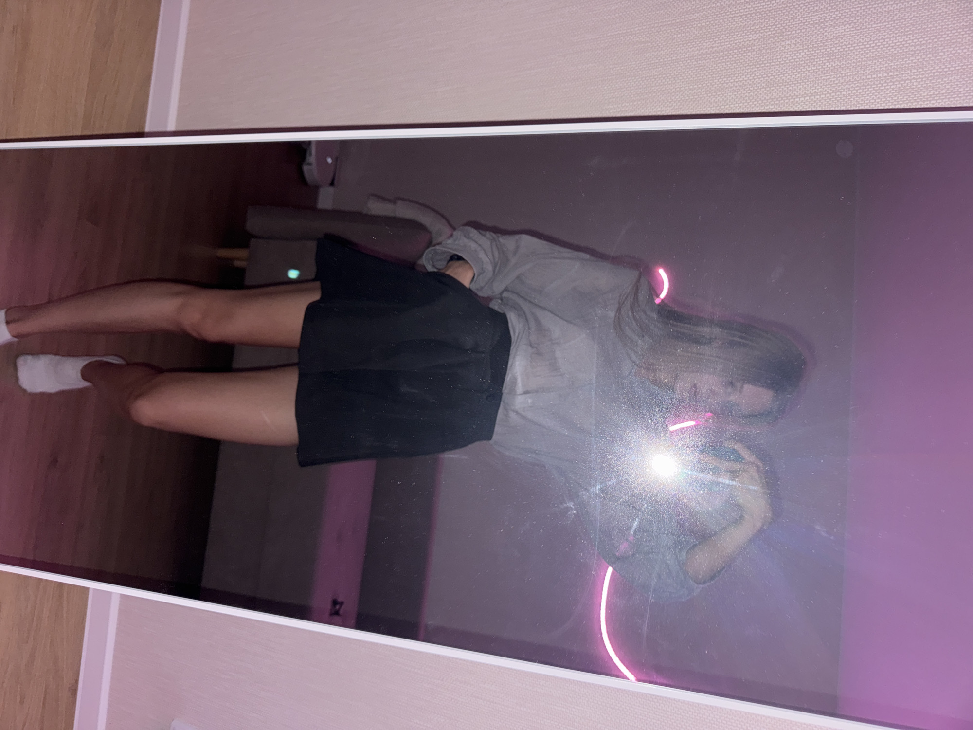 A full-body, slightly angled mirror selfie is captured in a room with pink and purple lighting. The subject, a young woman with long, light brown hair, stands facing the mirror, holding a phone with her right hand, the flash of which is illuminating the image. She is wearing a grey, oversized, long-sleeved top and a black mini-skirt. Her left hand is tucked into the pocket of her skirt. She has white socks and white sneakers on. The background in the mirror's reflection shows a couch, a neon pink curved light on the wall, and a robot vacuum cleaner on the floor. The room appears to be a bedroom or living area, with wooden flooring and pink-toned wallpaper on the left side of the frame. The image does not contain any specific information that would allow for its location to be determined as Perm, Russia.