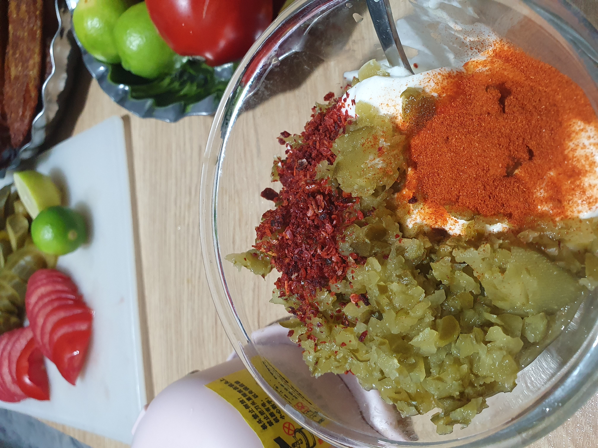 The image depicts a close-up view of a glass bowl containing a mixture of chopped green pickles, a dollop of white cream or yogurt, and a generous sprinkle of red chili flakes and paprika. A fork is partially visible, resting in the mixture. In the background, sliced tomatoes, limes, and a whole tomato are arranged on a white cutting board. Further in the background, a metal dish holds what appears to be grilled meat. A pink electric device with a yellow label containing Chinese text is also visible to the left. The scene is set on a wooden table. The image does not contain any information that would allow for its location to be specified in Kheyrabad, Iran.