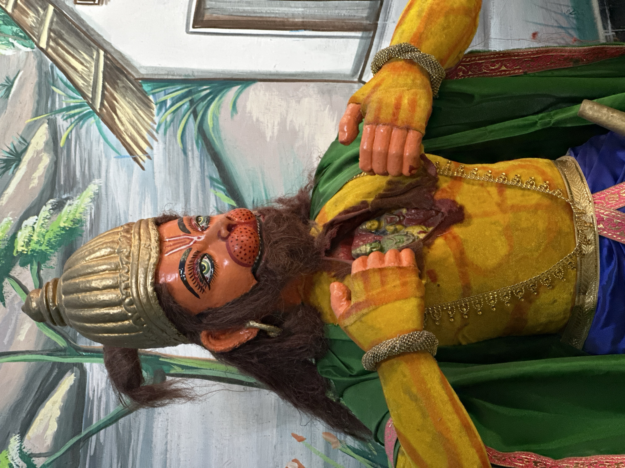 A statue of Hanuman stands against a painted background, likely in Varanasi, India. The statue is predominantly yellow and orange with green garments, and is depicted ripping open its chest to reveal an interior representation. A golden crown adorns its head. The painted background suggests an outdoor scene with structures and natural elements.