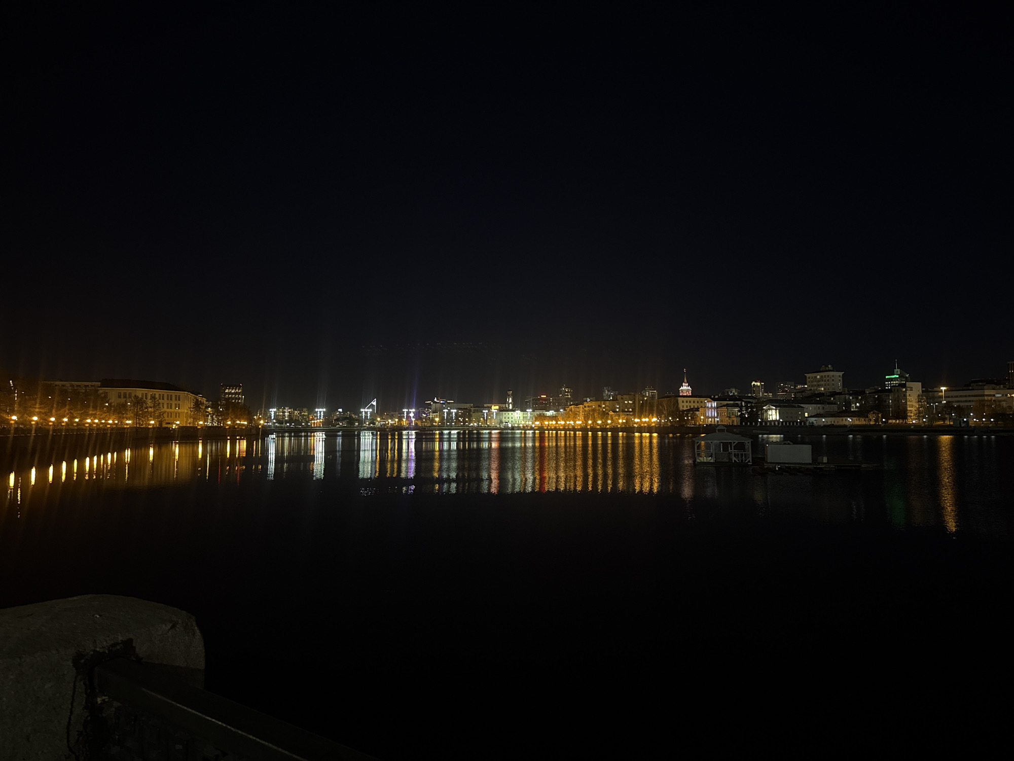 At night, the city of Yekaterinburg, Russia, is viewed across a dark body of water. The cityscape is lit up with various colors, and the lights are reflected in the water. In the foreground, a stone structure is visible.