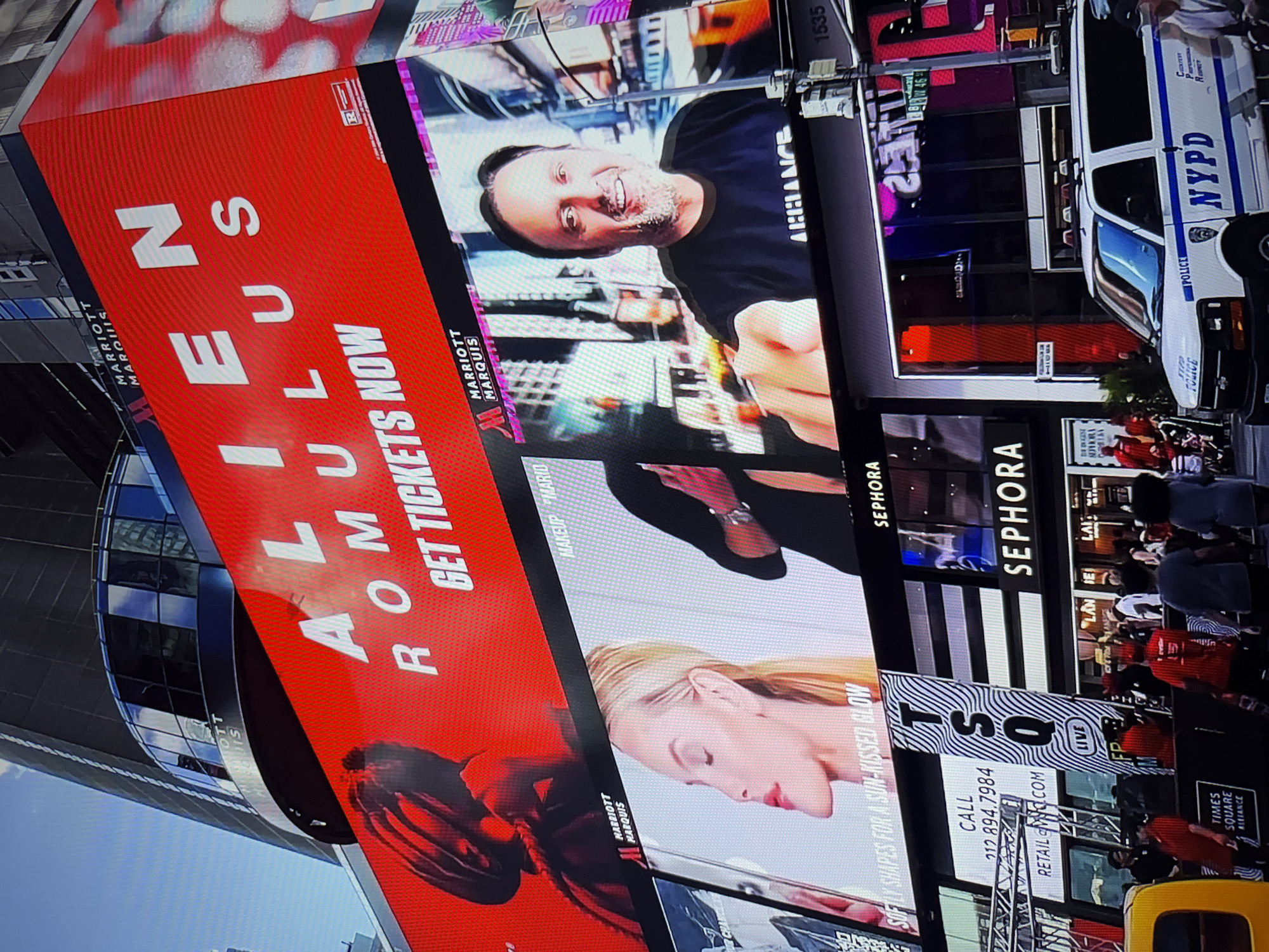 The image depicts a large LED billboard display in Times Square, New York City. The billboard prominently features the word "ALIEN" in large white letters against a red background, with "ROMULUS" below it and "GET TICKETS NOW" further down. Below this main display are smaller screens showing advertisements. One screen displays a woman's face with makeup, and another shows a smiling man. The scene also includes a police van with "NYPD" clearly visible on its side, and numerous people are visible in the background, suggesting a busy urban environment. The location is Times Square, New York City, not Burgas, Bulgaria.