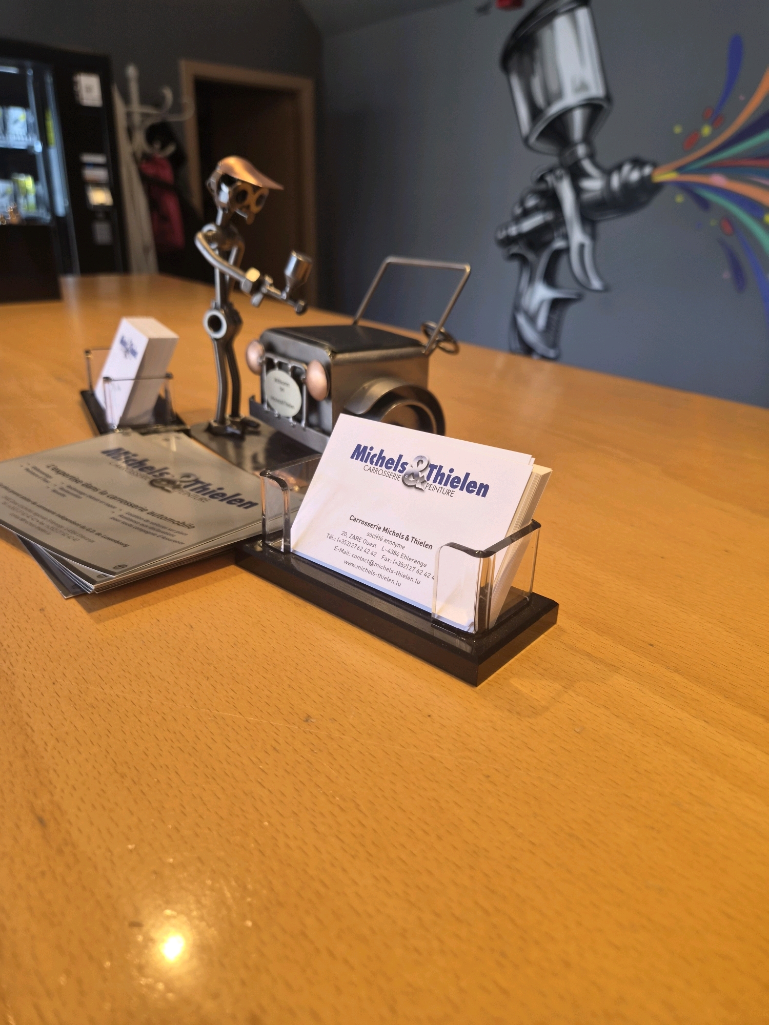 A metal figurine of a person holding a paint gun is standing next to a miniature metal car on a wooden table. To the left, a stack of brochures and business cards are displayed in a black organizer. A business card in the organizer reads "Michels & Thielen CARROSSERIE PEINTURE". The background shows a dark wall with a mural of a paint gun spraying colorful paint. The location is identified as Sanem, Luxembourg.