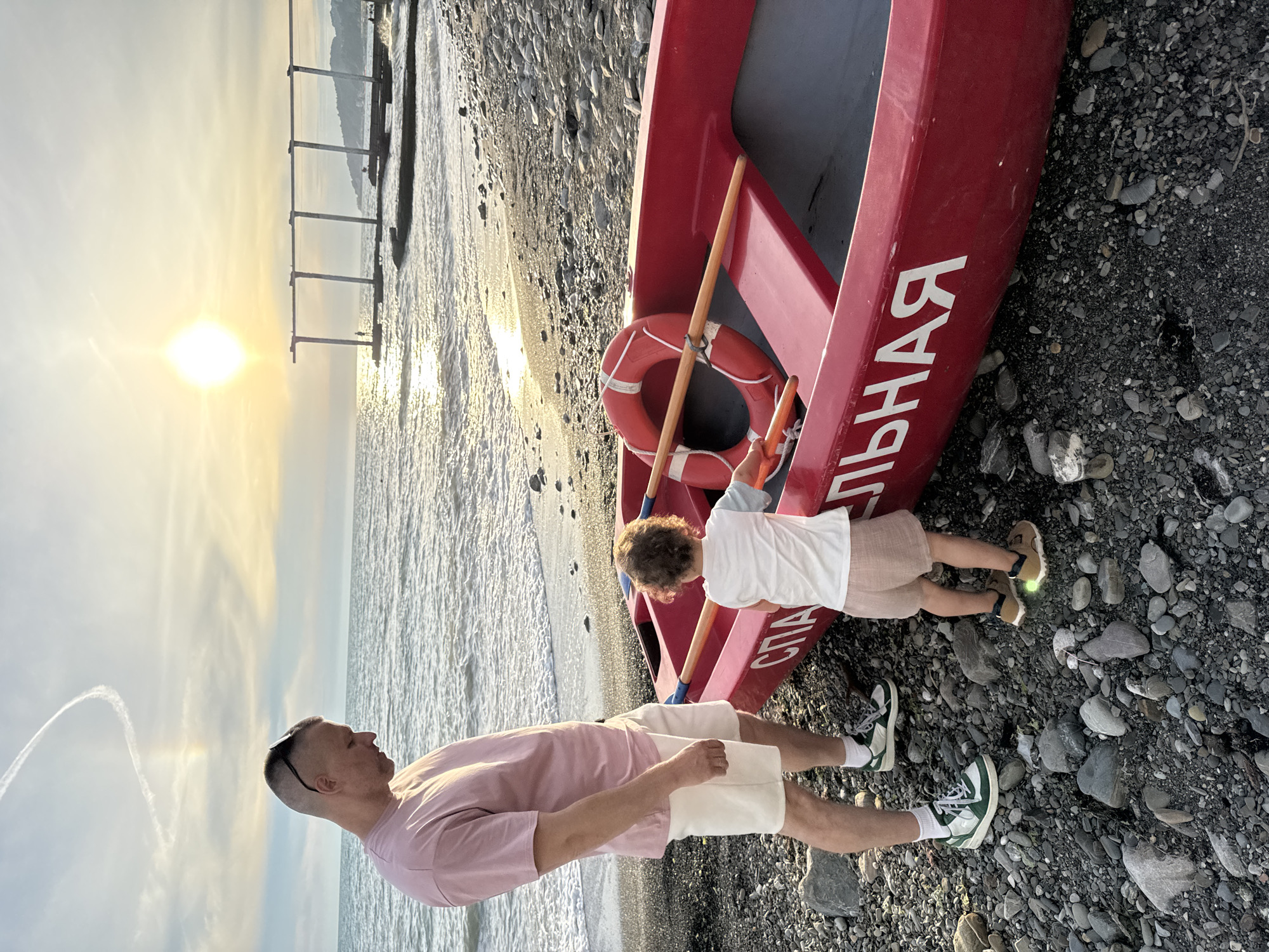 A man and a young child stand on a pebbled beach next to a red rowboat. The man, wearing a pink t-shirt and white shorts, looks down towards the boat. The child, in a white t-shirt and beige shorts, is facing the boat. The rowboat has the Russian word "СПАСАТЕЛЬНАЯ" (rescue) written in white letters along its side. A life preserver and oars are visible inside the boat. The sun is setting over the sea, casting a warm glow on the scene. In the background, a pier or structure extends into the water. The location is identified as Sochi, Russia.