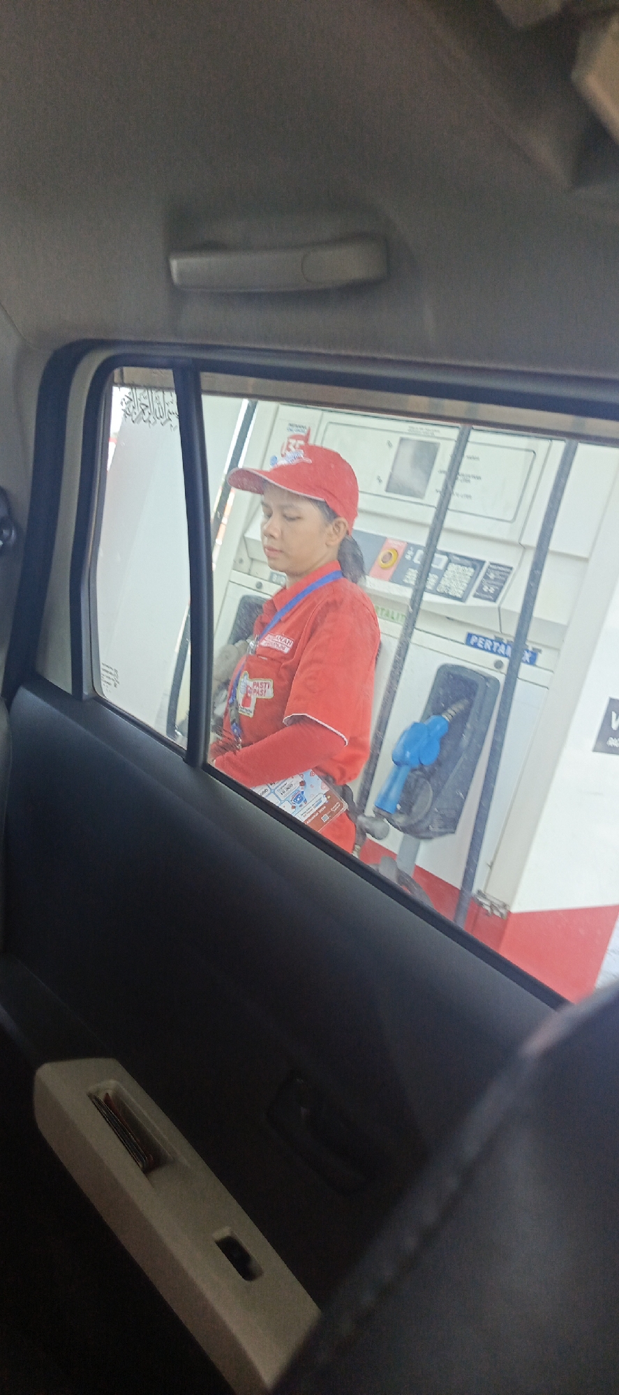 The image, taken from inside a vehicle in Sukaresmi, Indonesia, captures a female gas station attendant wearing a red uniform and a red cap. She is standing next to a gas pump, which is white with red accents and has a blue nozzle. The attendant appears to be looking downwards with her eyes closed or partially closed. The window of the vehicle obstructs a full view of her and the surrounding environment. Arabic calligraphy is visible on the window, and the interior of the vehicle, including a dark-colored dashboard and a light-colored pull handle, is also present in the foreground.