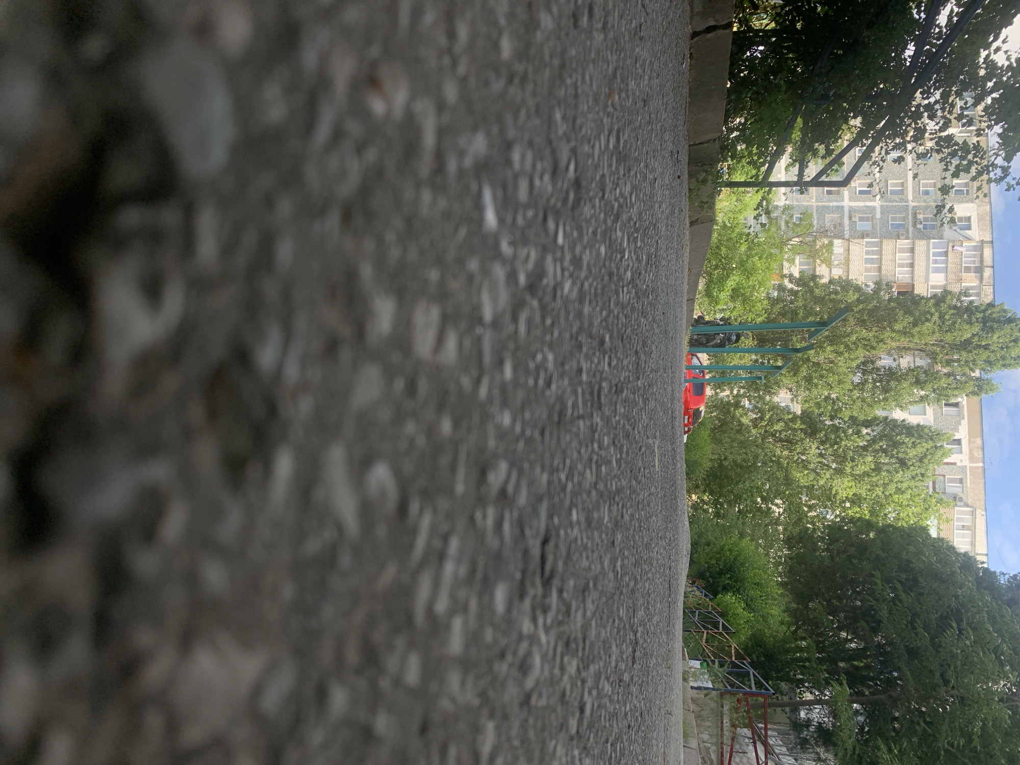 The image shows a low-angle view of an asphalt path leading upwards towards a residential building. On the left side of the path, there is a concrete barrier and lush green foliage. On the right side, a metal railing with red and blue accents runs alongside the path, which appears to be a set of stairs or a sloped walkway. In the background, a tall, multi-story apartment building with numerous windows stands against a bright sky with scattered clouds. Several mature trees with green foliage are scattered throughout the scene, partially obscuring the building and the sky. A red car is visible parked behind a green metal structure on the right side of the path. A motorcycle is parked near the green structure. The overall scene depicts a common urban environment with residential housing and natural elements. This location is likely in the city of Novorossiysk, Russia, judging by the architectural style of the building and the presence of mature trees in a temperate climate.