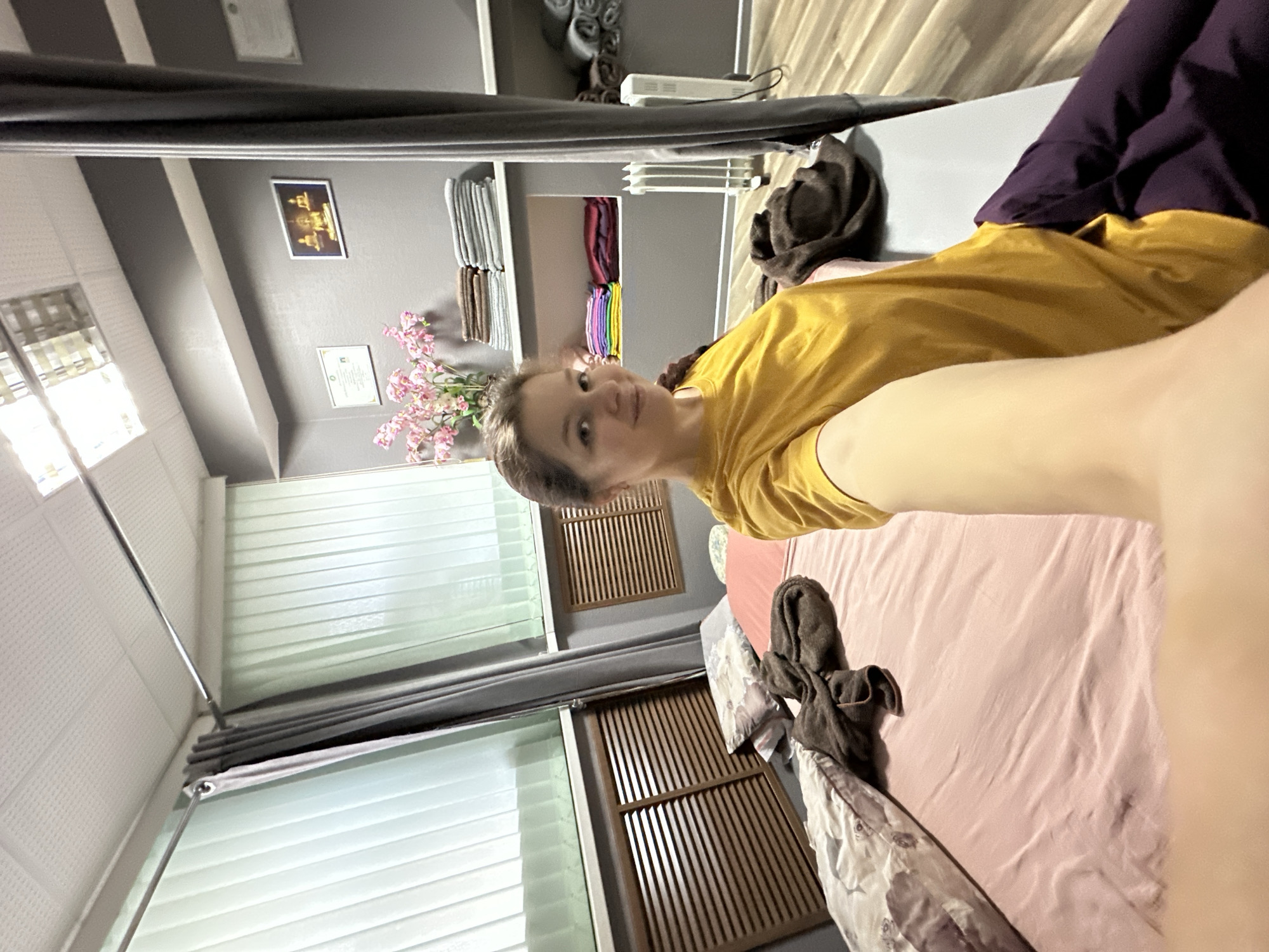 The image is a tilted, eye-level selfie captured by a woman lying down indoors, looking directly at the camera. The woman, with light hair (possibly in a braid) and wearing a mustard yellow short-sleeved top, has a calm, pleasant expression with a subtle smile. Her left arm is extended towards the camera, holding the device.
She is positioned on what appears to be a bed or massage table, covered with a light pink sheet. A dark brown folded towel rests near her head on the bed, and a dark purple garment is visible near her lower body. The room's background features grey walls with shelves displaying neatly folded towels in various shades of grey, brown, and a stack of colorful ones including red, purple, yellow, and green. An arrangement of artificial pink cherry blossoms adds a decorative touch.
On the wall, a framed picture, possibly depicting a golden Buddha or similar spiritual figure, hangs alongside another framed document or certificate, though the text on both is unreadable. The room has a mix of window coverings, including white horizontal blinds and brown wooden slatted panels. Dark grey curtains are drawn on a rod. A white rectangular device, possibly an air purifier or heater, is placed on one of the shelves. The ceiling features a textured or panelled design.
The overall scene suggests a wellness environment such as a massage salon, spa, or a dedicated relaxation room, indicated by the professional arrangement of towels and the general decor. The woman appears relaxed, perhaps enjoying a moment of rest. While the provided context suggests the location is Vladimir, Russia, the image itself does not contain specific visual cues to confirm this. The time of day is indeterminate due to the indoor setting and artificial lighting.