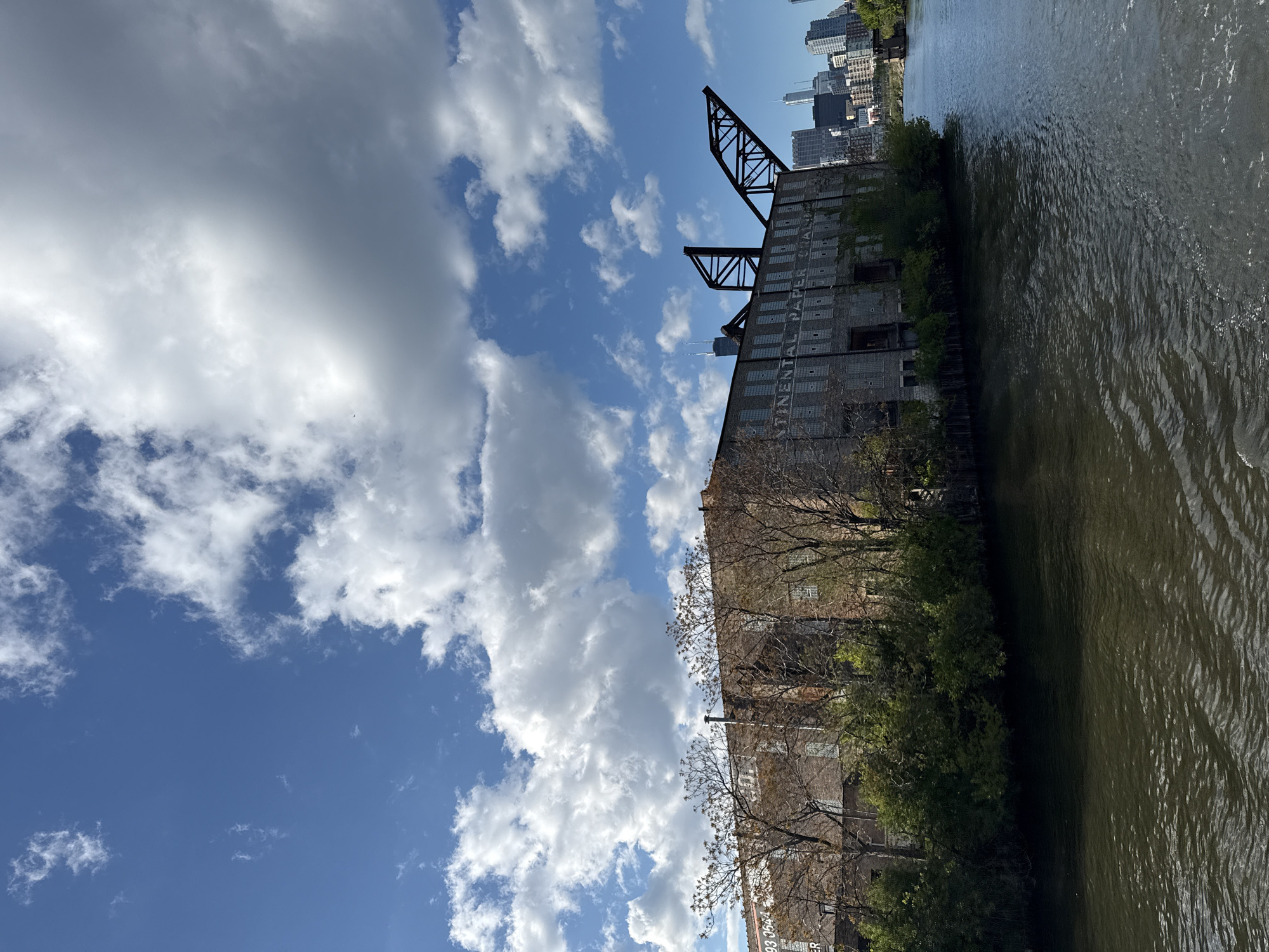 This image shows the Continental Paper Grading Company building along the Chicago River in Chicago, United States of America. The building is large and appears to be made of brick or stone, with multiple stories and a gantry crane mounted on the roof. The building's name is vertically written, though the full text is partially covered by leaves. Trees and bushes line the riverbank in front of the building. The Chicago skyline is visible in the background. The Chicago River is to the right of the building, and the sky has blue patches interspersed with large white clouds. The image appears to be taken from the water level.