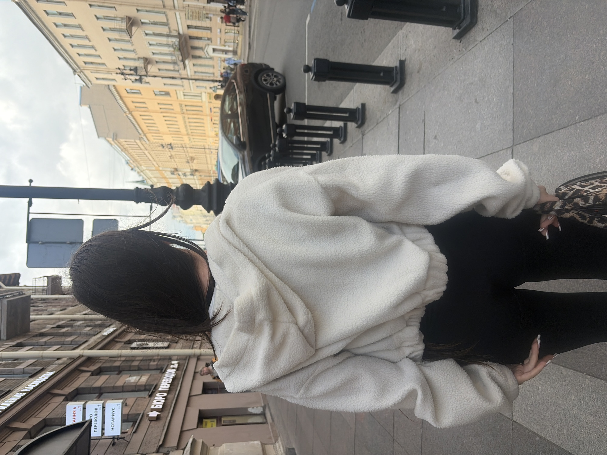 The image shows a woman from behind, wearing a white fleece sweater and black leggings. She is standing on a sidewalk with a patterned stone pavement. To her right, there is a black metal bollard. Behind her, there is a street with a dark-colored car and more bollards. In the background, there are buildings with classical architecture, characteristic of Saint Petersburg, Russia. The sky is overcast, suggesting it might be a cloudy day. The buildings have many windows and some signs. One sign, partially visible on a building to the left, appears to be in Cyrillic script. Another sign, clearer, reads "БЮРО ПЕРЕВОДОВ" which translates to "Translation Bureau". The overall impression is a candid shot of someone on a city street.