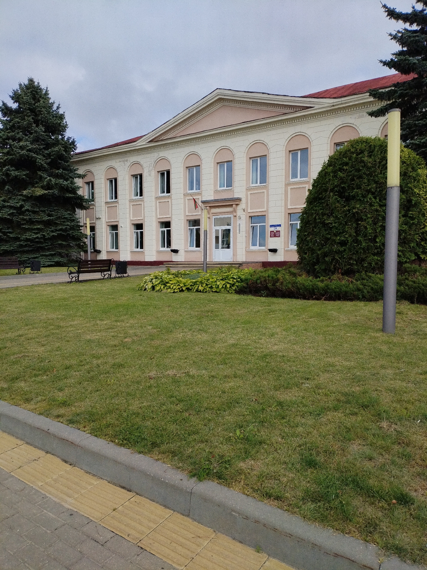 The image shows a two-story building in Bialynichy, Belarus, with a light pink facade and arched windows. A well-maintained lawn surrounds the building, with a sidewalk and curb in the foreground. A tall evergreen tree stands to the left of the building, and a smaller, dense shrub is to the right. Two dark benches with trash receptacles are placed on the lawn. A yellow lamppost is visible near the shrub. A red-tiled roof caps the building. The sky is overcast, suggesting a cloudy day.