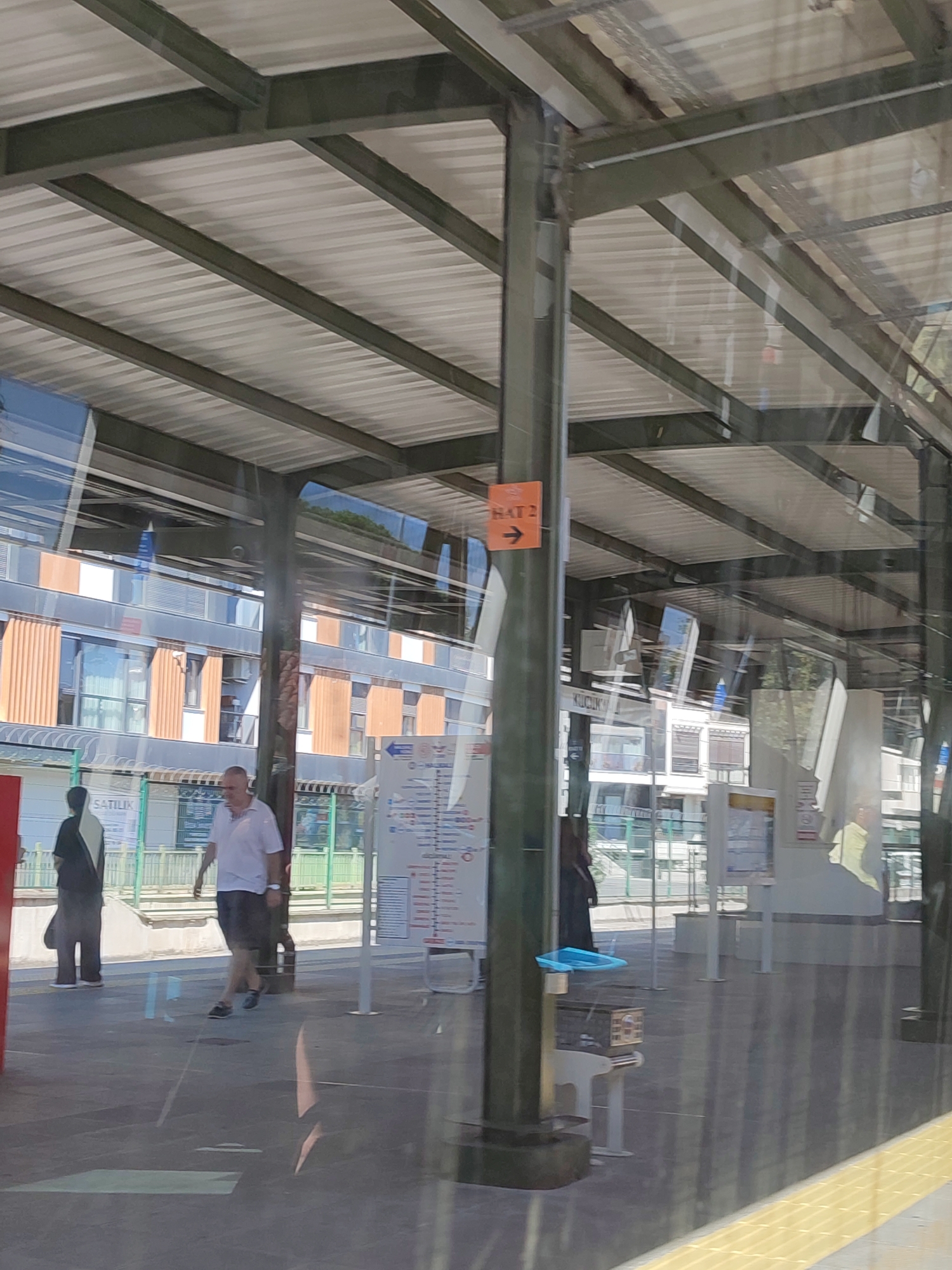 The image depicts a view through a glass enclosure, likely a train station or public transport waiting area, overlooking an outdoor platform. A modern building with a facade of orange vertical panels and windows is visible in the background, suggesting an urban setting. A man in a white polo shirt and dark shorts is walking on the platform, while another person in a black outfit stands to his left. The platform is covered by a corrugated metal roof supported by dark metal beams and pillars, typical of an open-air transit structure. A sign with orange background and black text pointing to the right with the text "HAT 2" is affixed to one of the pillars. The ground is paved with dark tiles. Reflections in the glass indicate the presence of other structures and possibly the sun. The scene suggests a sunny day. The presence of the sign "HAT 2" and the architectural style of the building are consistent with a location in Maltepe, Turkey, a district known for its modern infrastructure and transportation hubs.