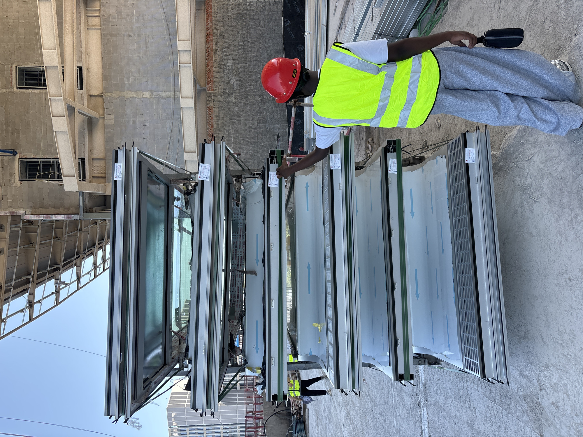 In Liangjiang, China, a construction worker wearing a red hard hat and a bright yellow safety vest stands in front of a stack of large, framed glass panels on a construction site. The worker is holding a black bag in their right hand. The panels are stacked on a metal frame, with some appearing to be windows or doors. In the background, a building under construction is visible, along with scaffolding and other construction materials. The sky is clear and blue.