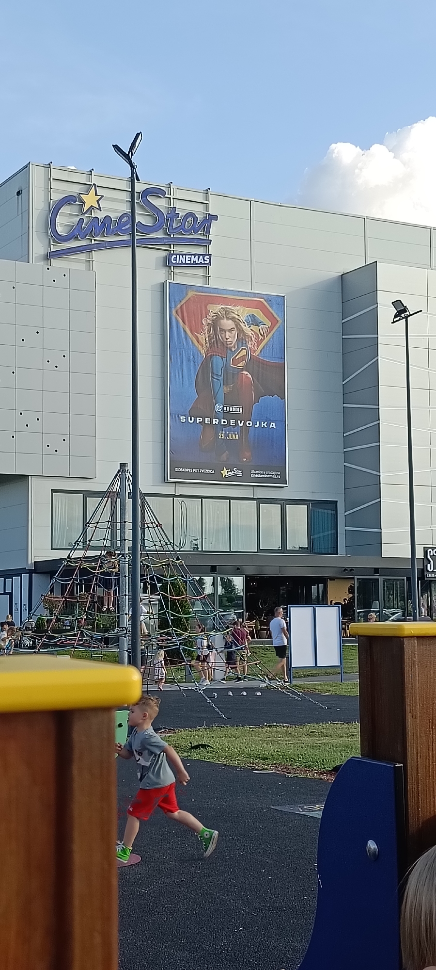 The image shows an exterior view of a CineStar cinema complex on a sunny day. In the foreground, a young boy in a grey t-shirt and red shorts is running on a black playground surface, with a yellow and brown play structure partially obscuring him. Behind him, a large rope climbing structure is visible, with several children and adults on and around it.
The cinema building is a modern structure with large windows and a prominent "CineStar CINEMAS" sign. A large movie poster is displayed on the side of the building, featuring the DC Comics character Supergirl. Text on the poster reads "STUDIOS SUPERDEVOJKA 25. JUNA" (Supergirl June 25th) and mentions "CINEMAS PET ZVEZDICA" (Cinemas Five Stars) and "Ulaznice u prodaji na cinestarcinemas.rs" (Tickets on sale at cinestarcinemas.rs). The sky is blue with some scattered white clouds, suggesting fair weather. The scene appears to be a recreational area, likely a shopping mall or entertainment complex, in Novi Sad, Serbia.