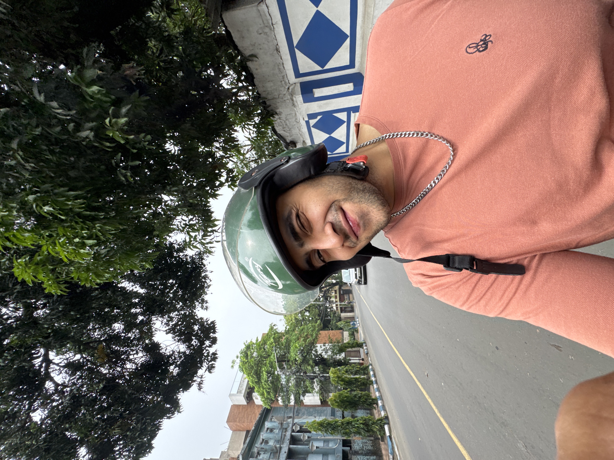 This portrait-oriented image, rotated 90 degrees clockwise, captures a close-up selfie of a smiling young man in an outdoor urban setting. The subject wears a green motorcycle helmet, on which the brand "VEGA" is distinctly visible. He is dressed in a peach or salmon-colored t-shirt featuring a small, dark blue embroidered logo on the left chest, which appears to be stylized initials like "GS" or similar. A thick silver chain is visible around his neck. His expression is one of contentment, with a clear smile on his face.
The background details confirm a daytime scene under an overcast sky. Lush green trees are prominent to his left (appearing at the top of the rotated image). To his right, a paved road with a yellow dividing line stretches into the distance, with vehicles barely visible further down. The setting includes various buildings and, notably, a white wall adorned with a repeating pattern of blue diamond shapes, a distinctive architectural feature often seen in Kolkata, India, aligning with the provided location context. The presence of the helmet and the road strongly suggest he is on a motorcycle or scooter. The overall impression is a casual moment captured while traveling.