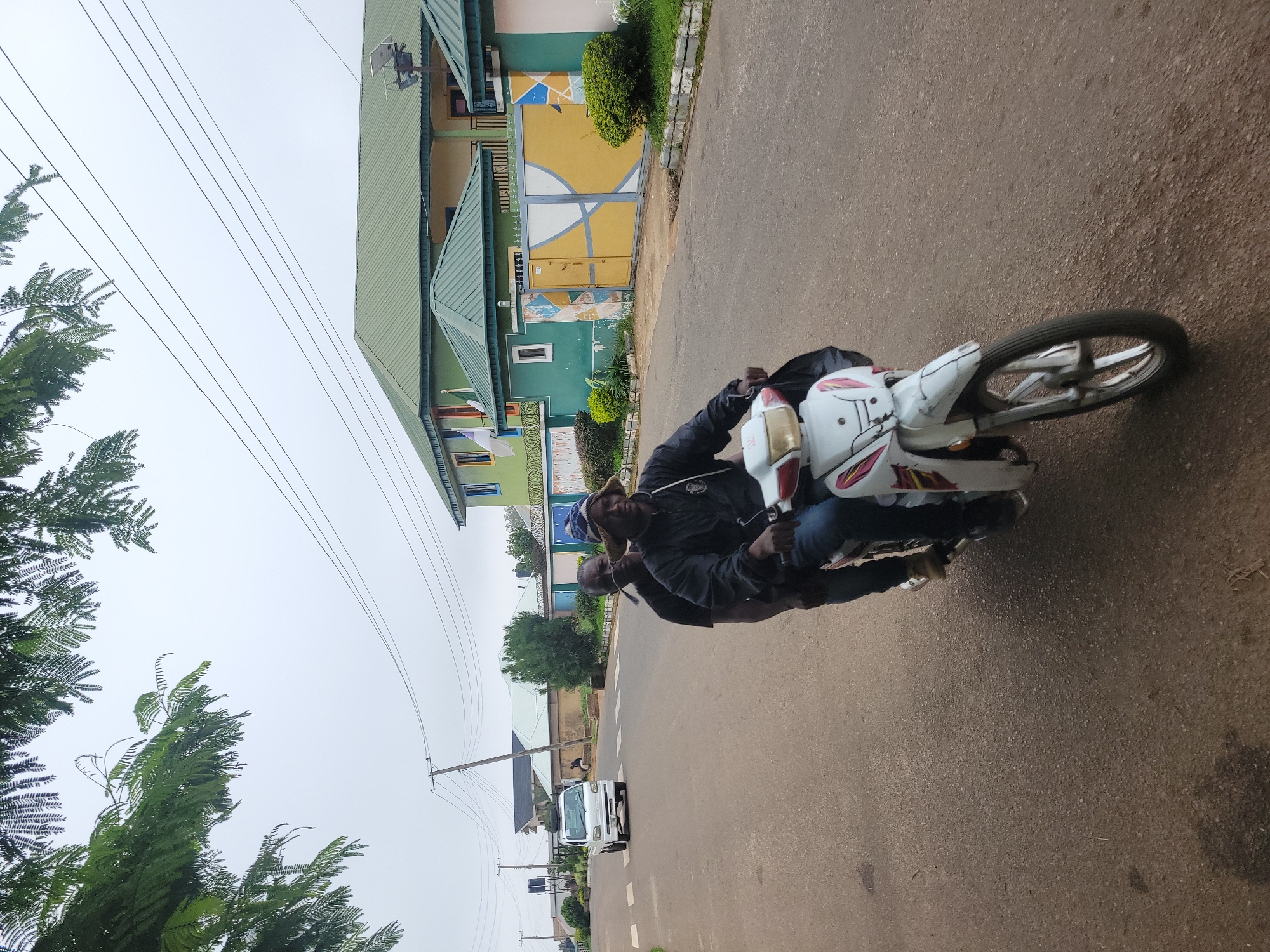 Two men ride a white motorcycle down a paved road in Jos, Nigeria. The man in front, wearing a dark jacket and a blue and white patterned hat, is driving. The man behind him, wearing a dark shirt, is looking forward. The motorcycle is white with red and yellow accents. The road is lined with houses and trees. A white truck is visible in the background, driving away from the motorcycle. The sky is overcast.
