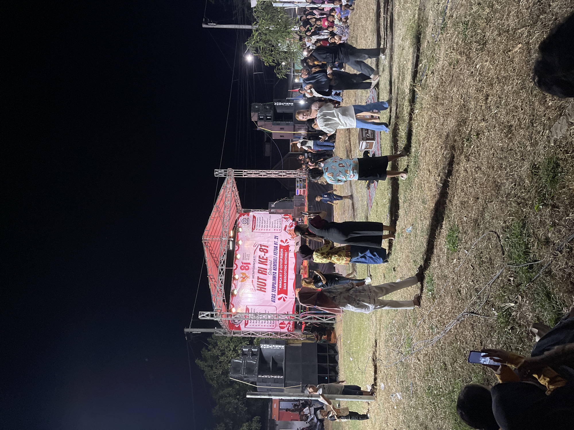 In Jatirembe, Indonesia, at nighttime, a large outdoor stage is set up for an event. A banner on the stage reads "HUT RI KE-81 SELAMAT" and lists various names. Several people are on the stage, seemingly performing or participating in the event. In front of the stage, a crowd of people are gathered on a grassy field. Some individuals are standing and watching the stage, while others are sitting. To the left of the stage, large speaker systems are set up. The environment is illuminated by stage lighting and some ambient light from nearby structures and lamps. The overall scene depicts a lively community event taking place in an outdoor urban setting.