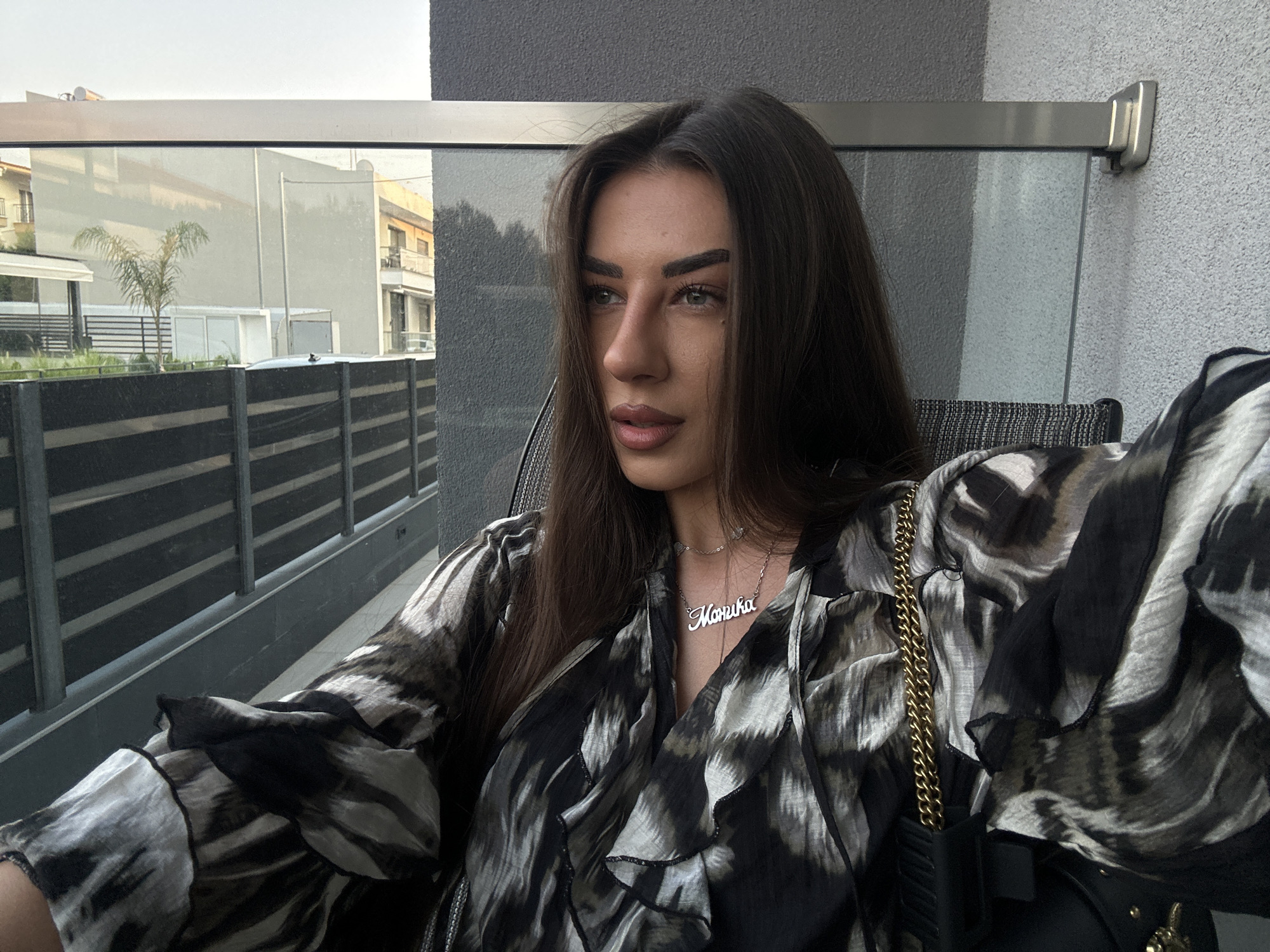 A medium shot captures a young woman with long dark hair, wearing a black and white patterned blouse and a gold necklace with a nameplate, sitting outdoors. The woman's gaze is directed to her left. Behind her, a modern building with balconies and a glass railing is visible, along with a black fence. In the distance, trees and other buildings can be seen. The setting appears to be a balcony or patio area. The location is Ofrynio Beach, Greece.