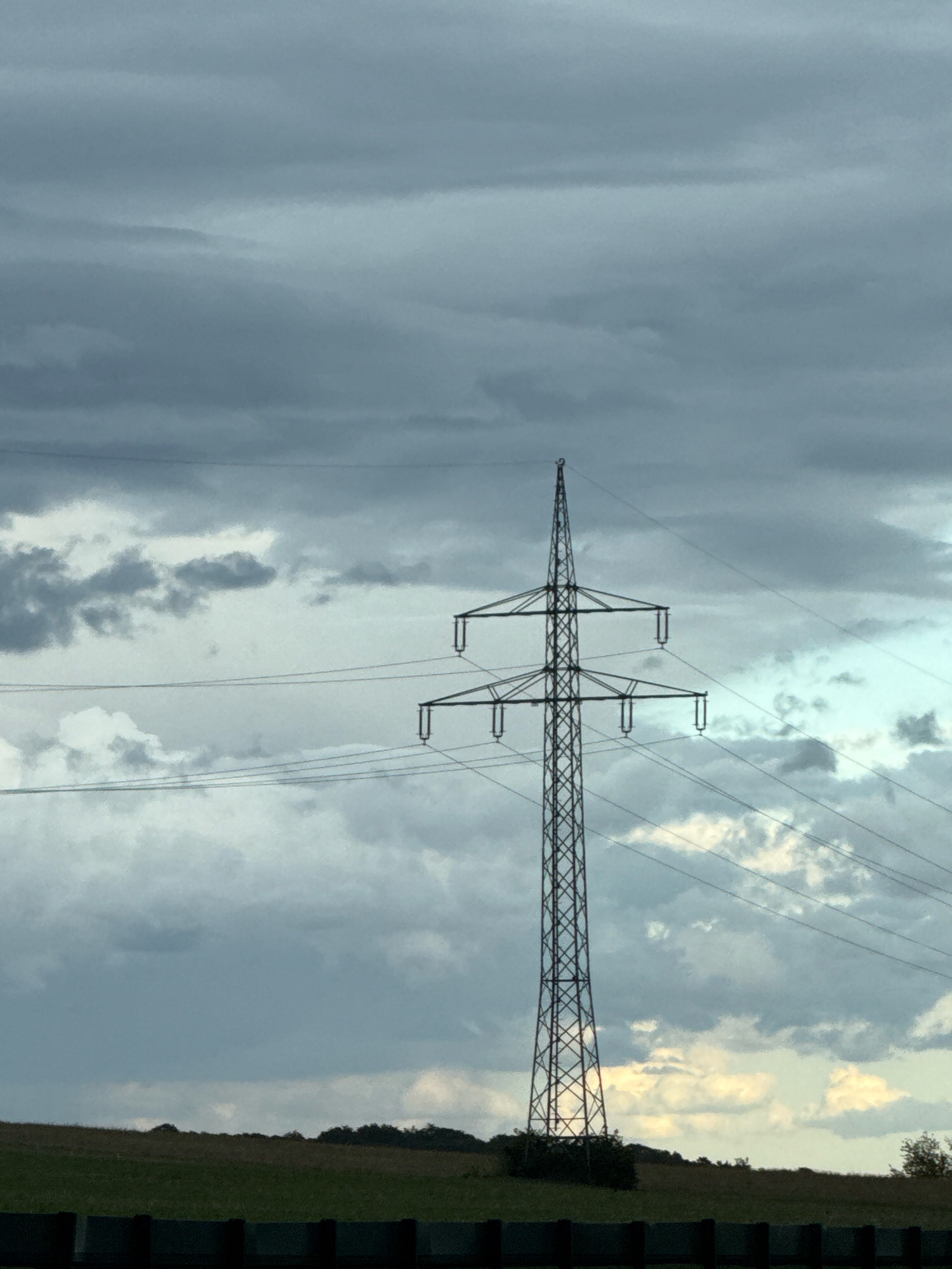 The image shows a tall, lattice-style electrical pylon set against a cloudy sky. The pylon is in the foreground, dominating the center of the frame, with its wires extending out of the frame to the left and right. Below the pylon, a field is visible, with a line of trees on the horizon. The sky is filled with a mix of dark, heavy clouds and lighter, scattered clouds, suggesting an overcast or approaching storm. The overall mood is somewhat dramatic due to the sky conditions. The image appears to be taken from ground level, looking upwards towards the pylon and the sky. The location is identified as Bad Lauterberg im Harz, Germany.
