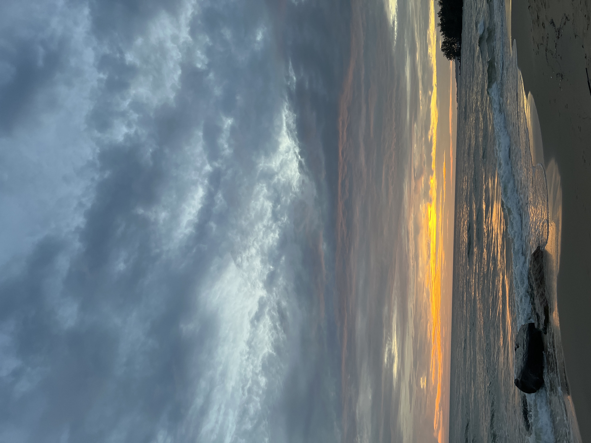 The image depicts a serene beach scene at sunset in Phú Quốc, Vietnam. The sky is filled with dramatic, textured clouds in shades of gray, blue, and hints of pink and orange from the setting sun. The ocean is calm, with gentle waves lapping at the sandy shore. In the foreground, a large dark rock sits partially submerged in the shallow water, accompanied by smaller rocks on the beach. The sand is wet and reflects the warm glow of the sunset. To the far right, the silhouette of a lush green tree line suggests the presence of land. The overall atmosphere is peaceful and tranquil, capturing the natural beauty of a coastal sunset.